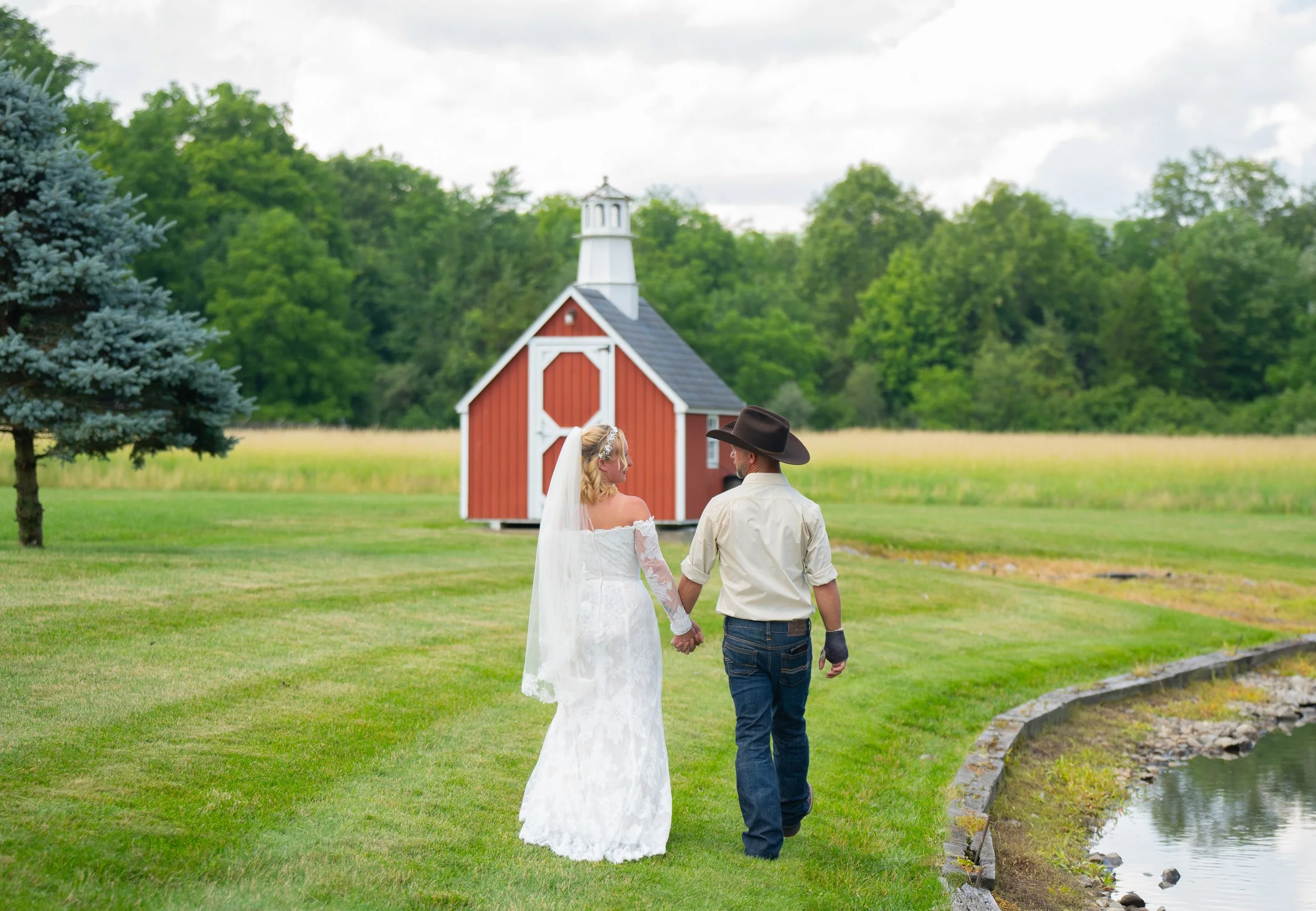 Bride and groom walking towards a red barn on a grassy field, with a pond and trees in the background.