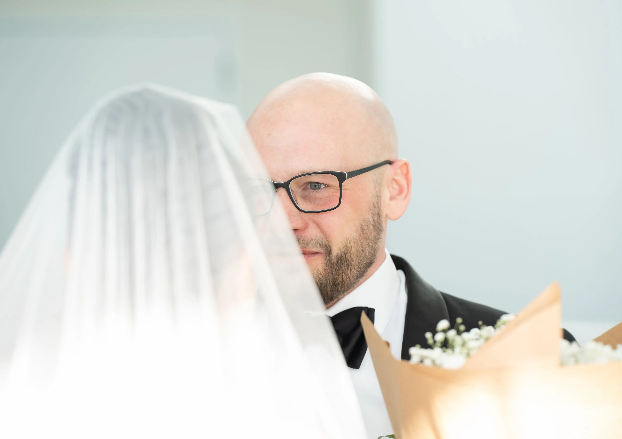 Bride and groom during wedding ceremony, bride veiled, groom in suit with glasses holding flowers.