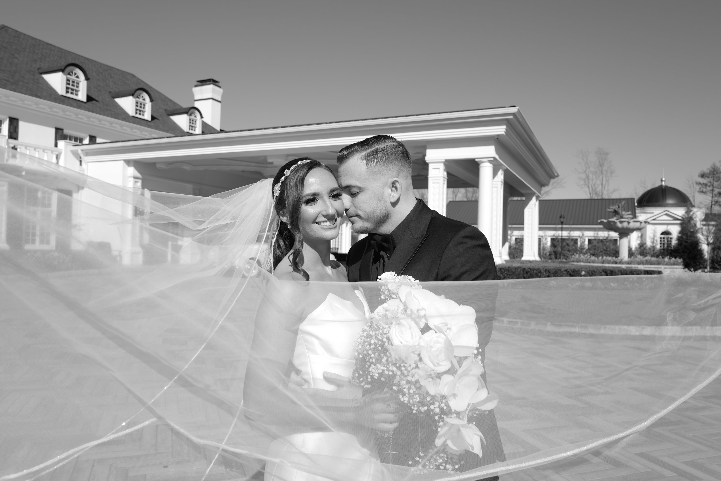 Black and white wedding photo of a couple embracing with a veil and floral bouquet in front of a large building.