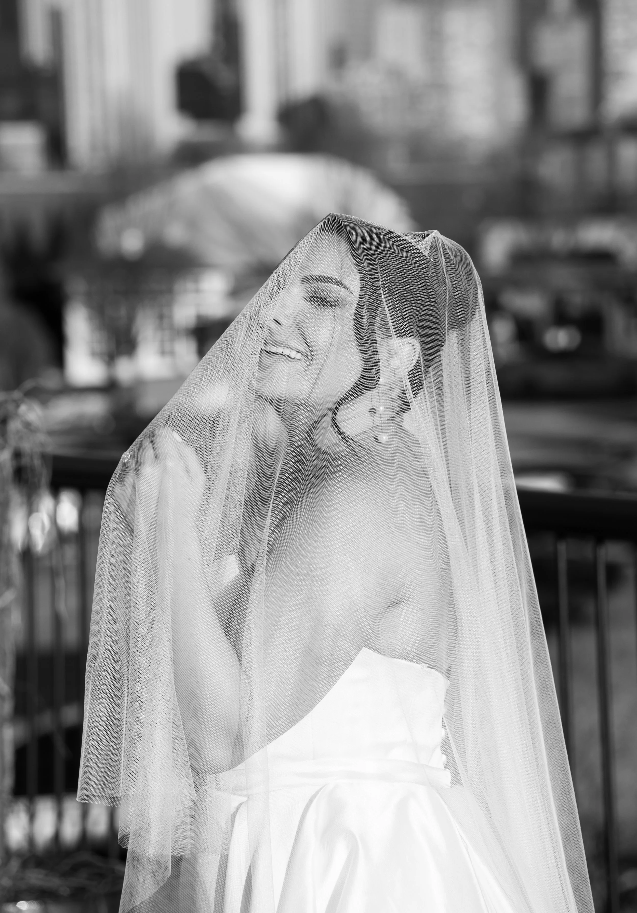 Black and white photo of a smiling bride wearing a veil and wedding dress, standing outdoors.