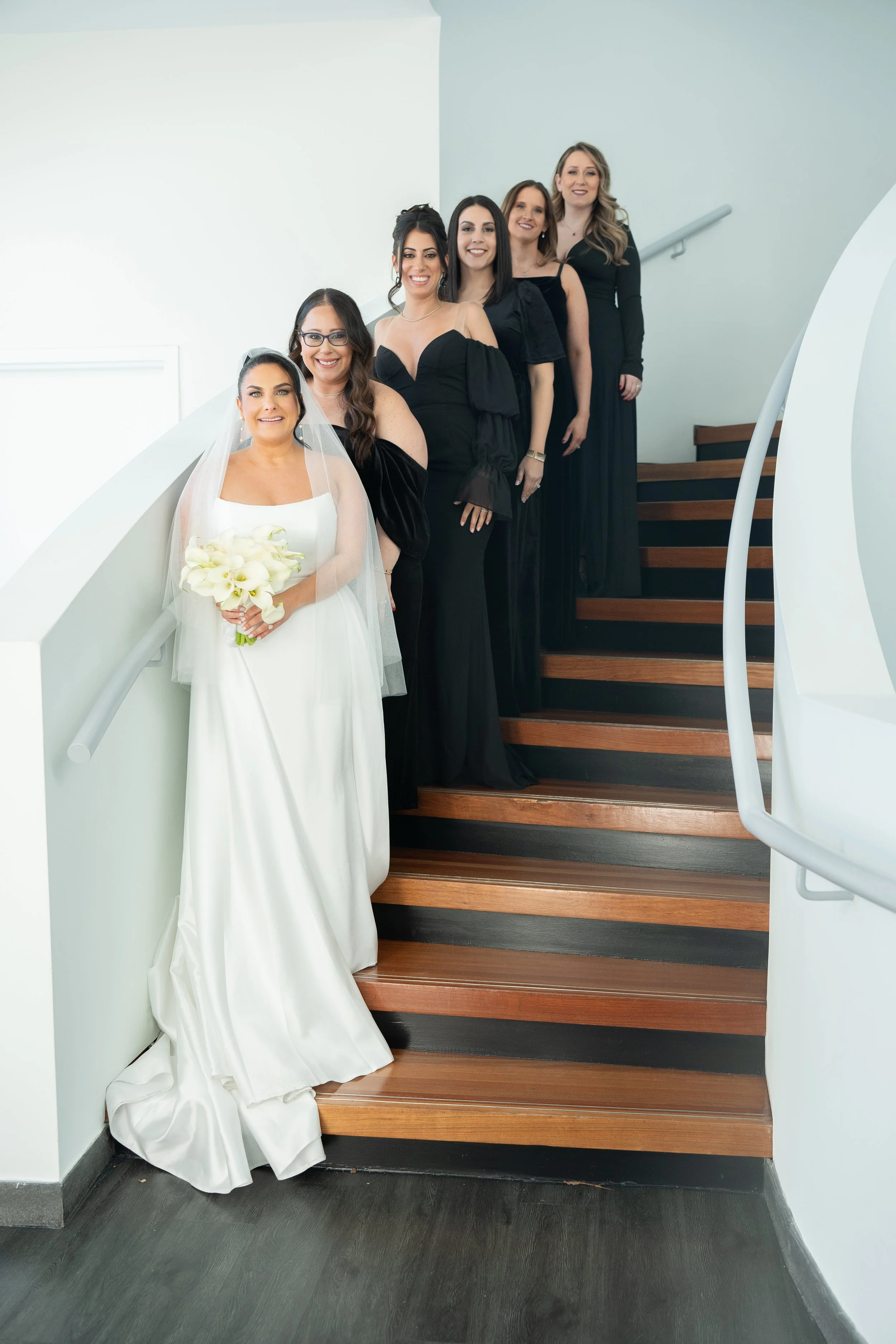 Bride in white gown with bridesmaids in black dresses on curved staircase.
