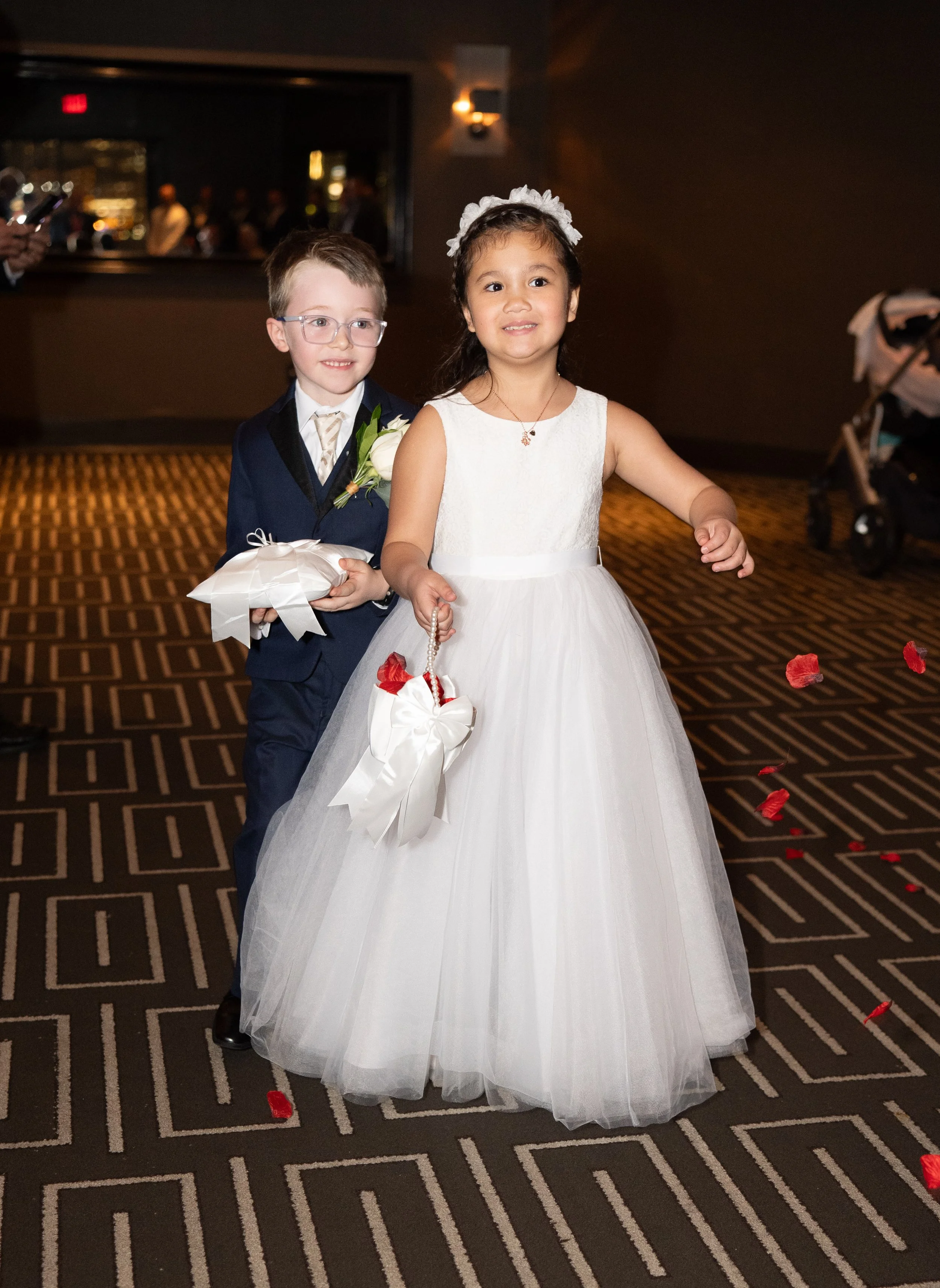 Two children walking down an aisle, a boy in a suit holding a pillow, and a girl in a white dress scattering petals.