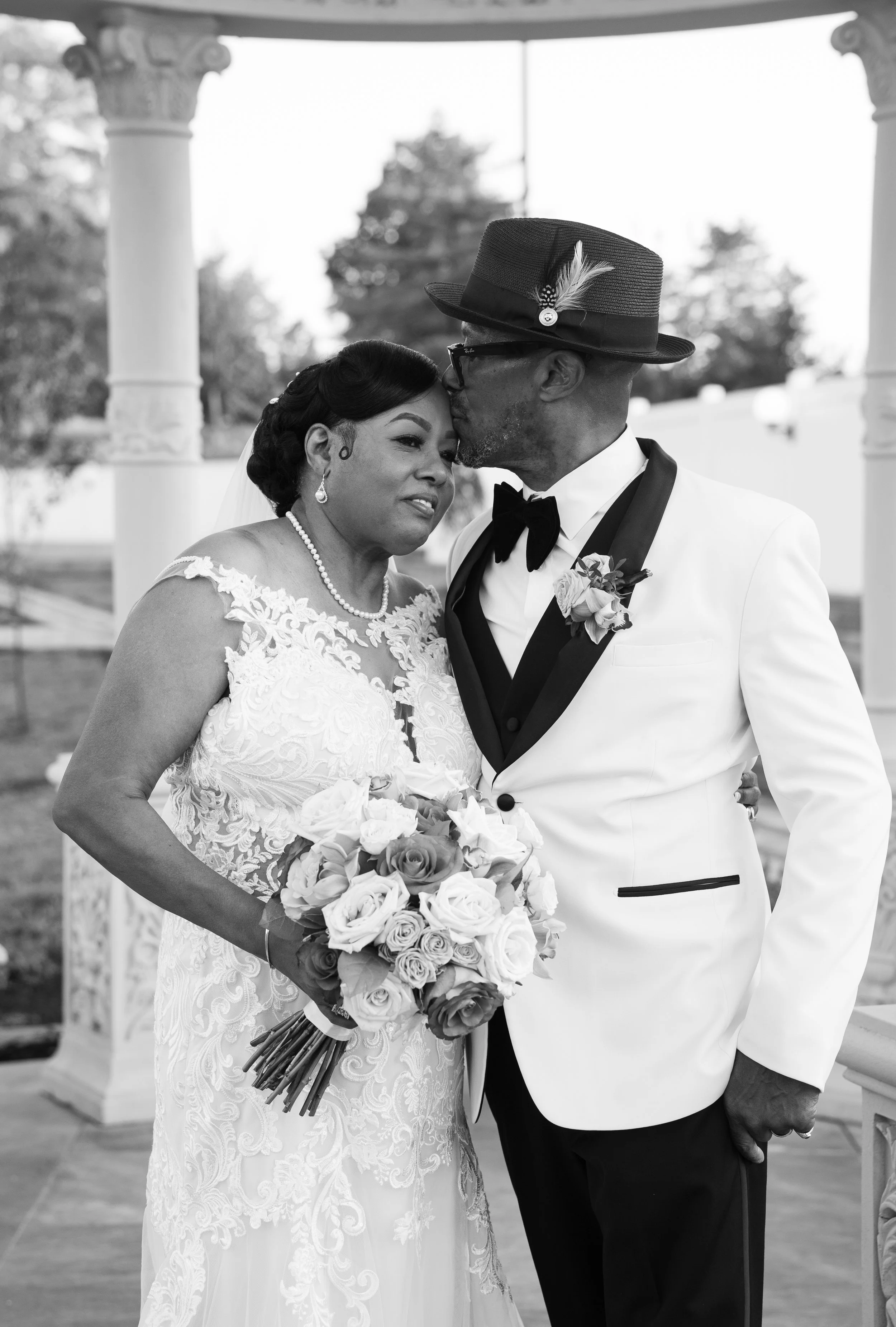 Black and white photo of a bride and groom embracing in wedding attire under a gazebo.