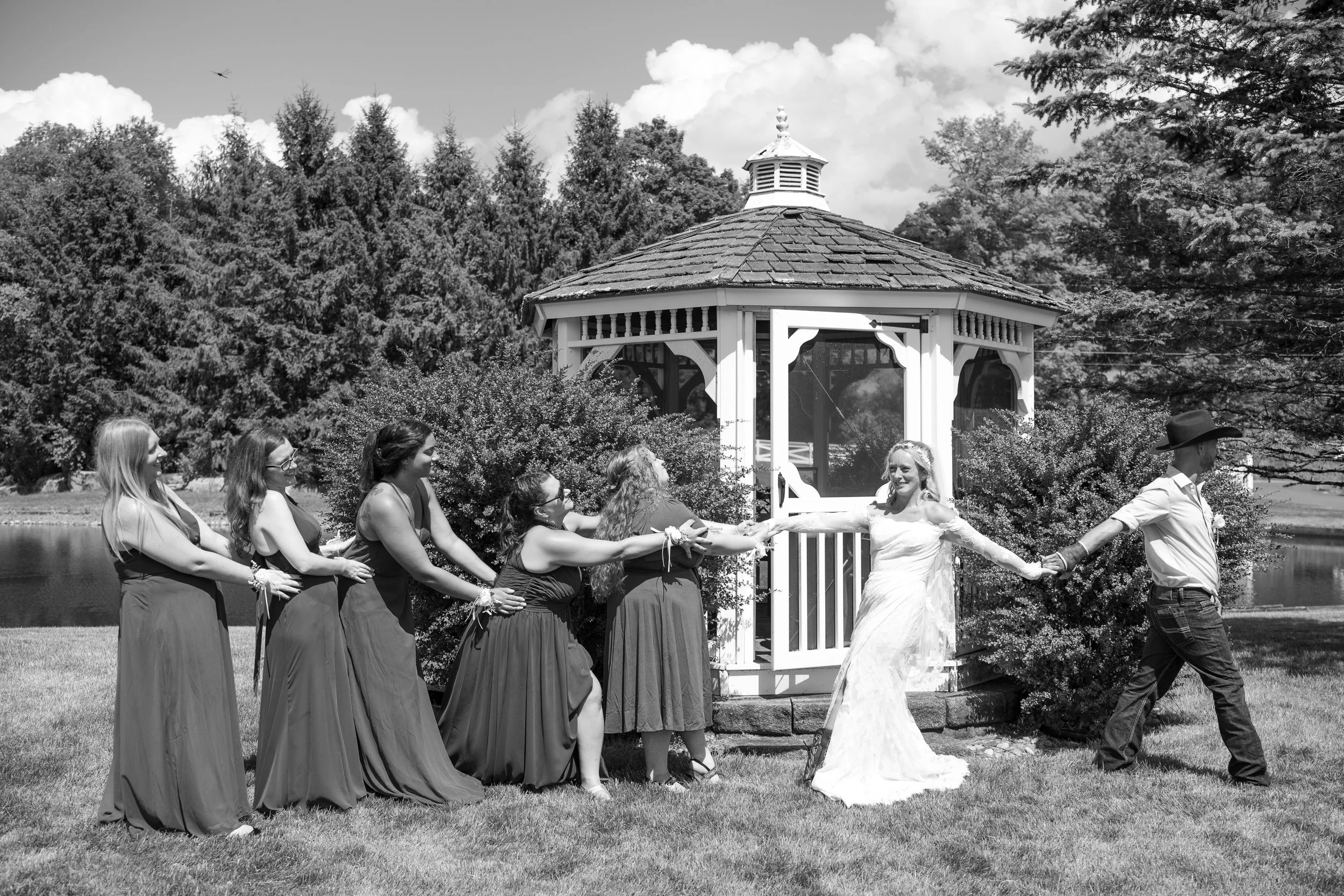 Bride in wedding dress being playfully pulled by bridesmaids in formal dresses and a man in a cowboy hat by a gazebo.