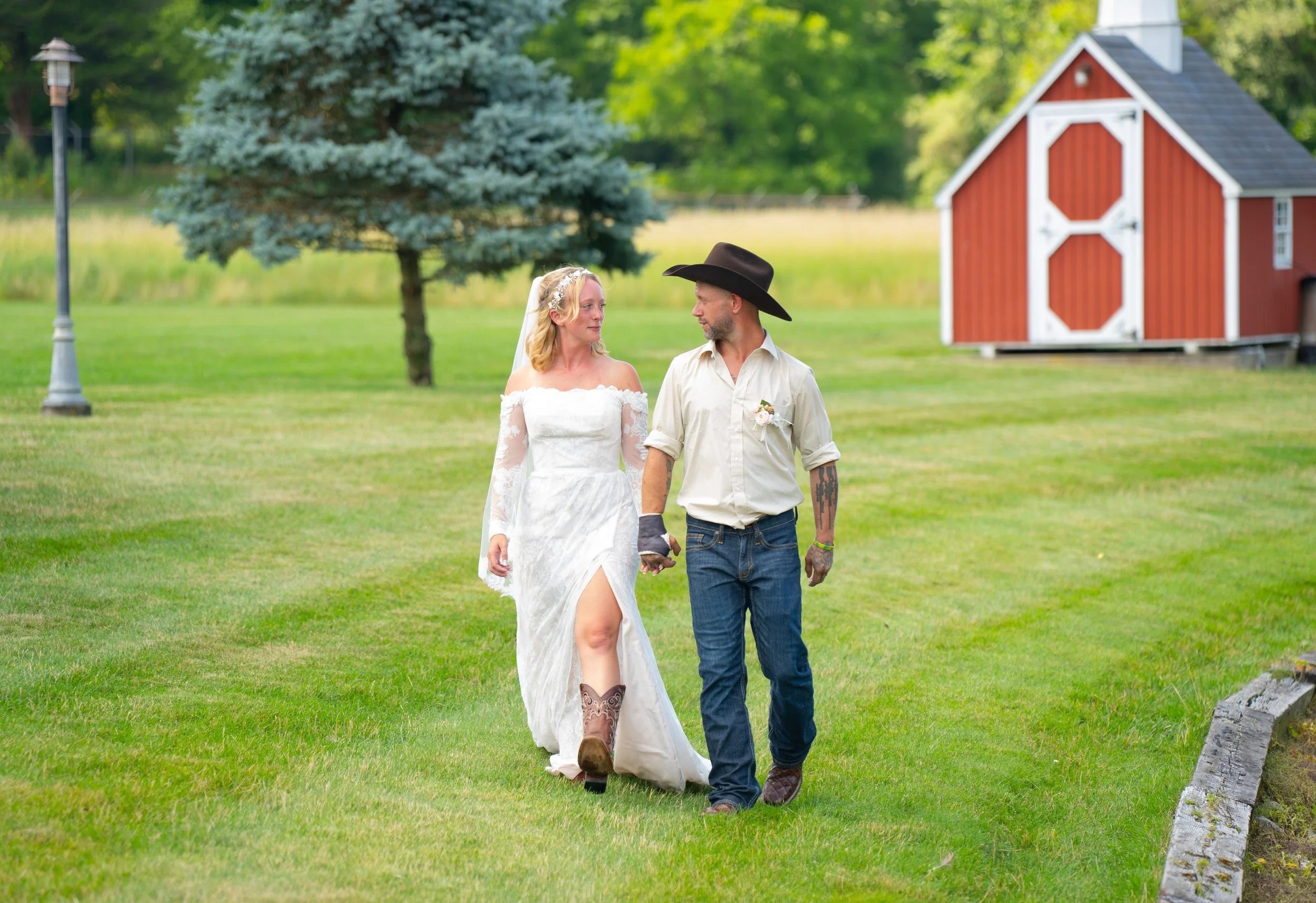 Bride and groom walking on grass near a barn, wearing cowboy attire, outdoors.