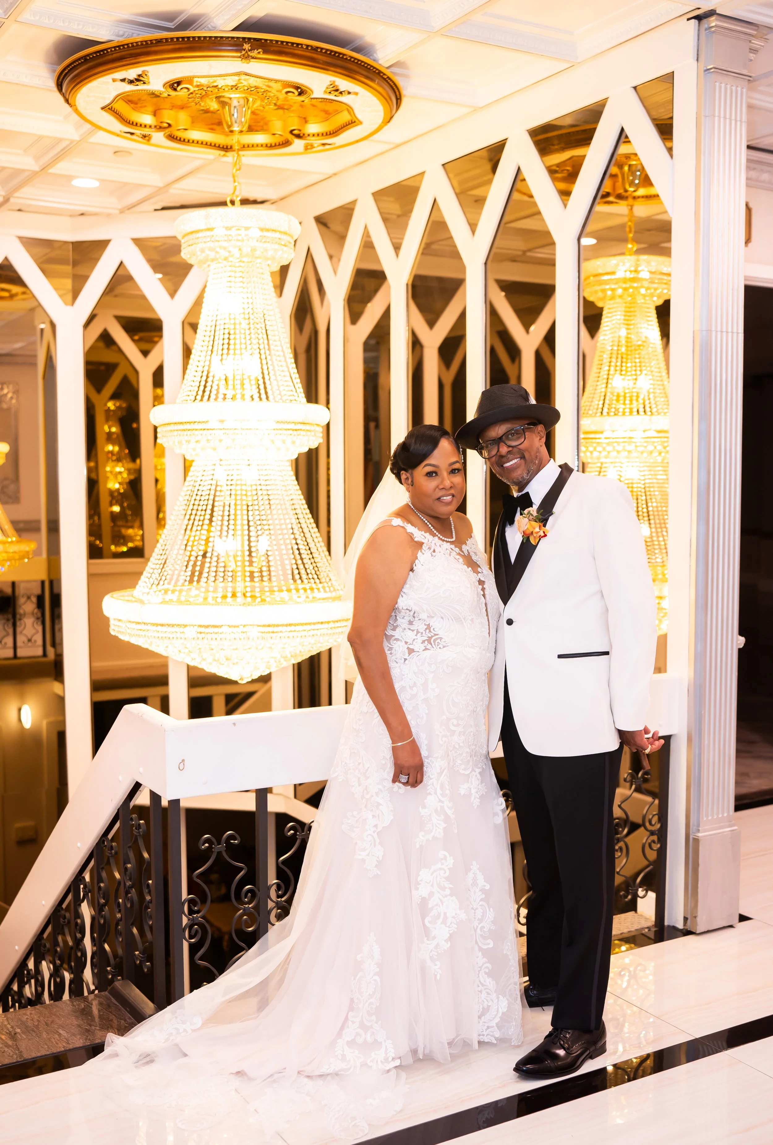 Bride and groom standing on a staircase with elegant chandeliers in the background; the bride wears a white lace gown and the groom a white tuxedo with a black hat.