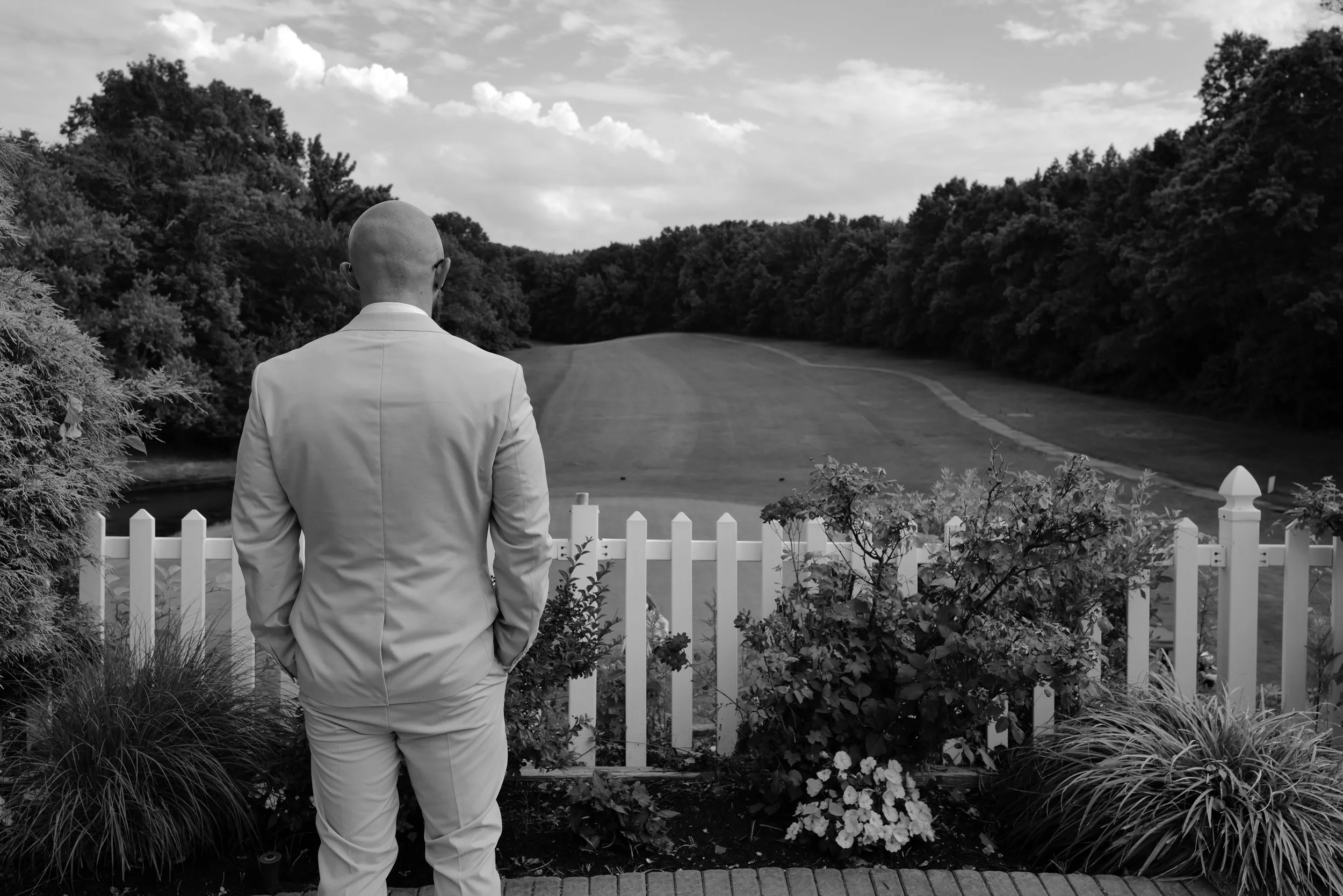 Man in suit facing a golf course, standing near a white picket fence with surrounding greenery.