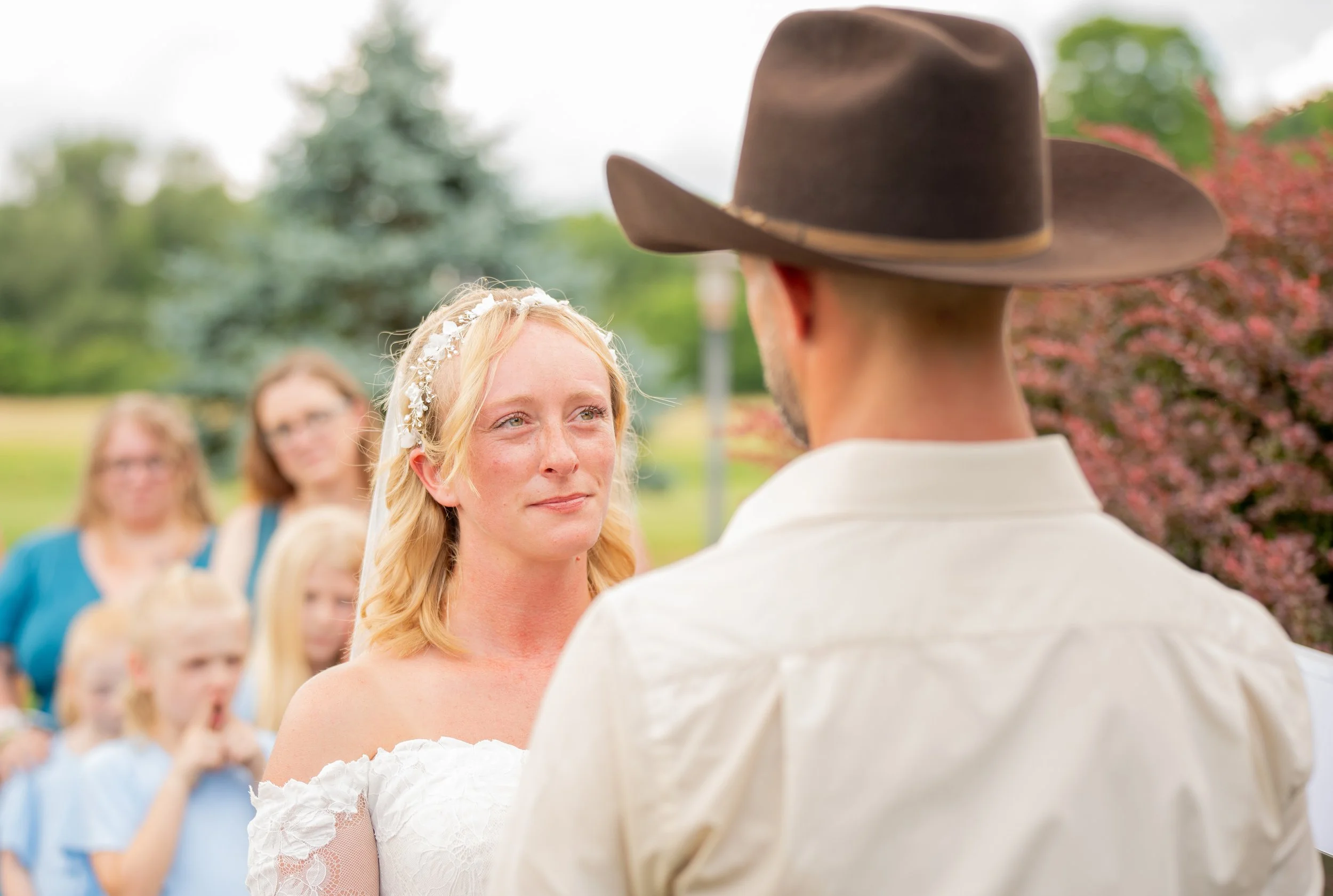Bride and groom exchanging vows outdoors, groom wearing cowboy hat, audience watching in background.