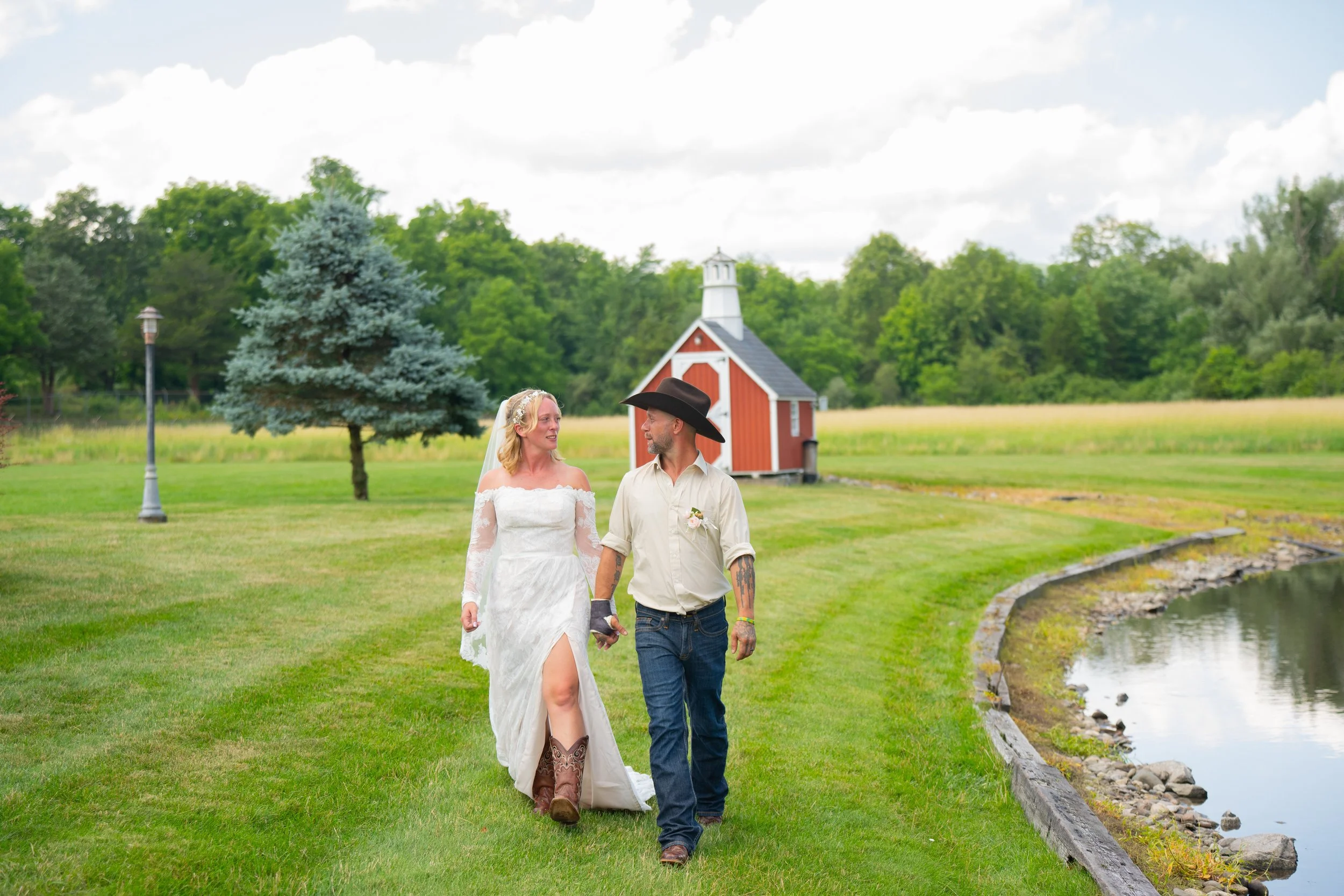 Bride in white dress and groom in cowboy hat walking outside, near a pond and red barn, with green trees in the background.