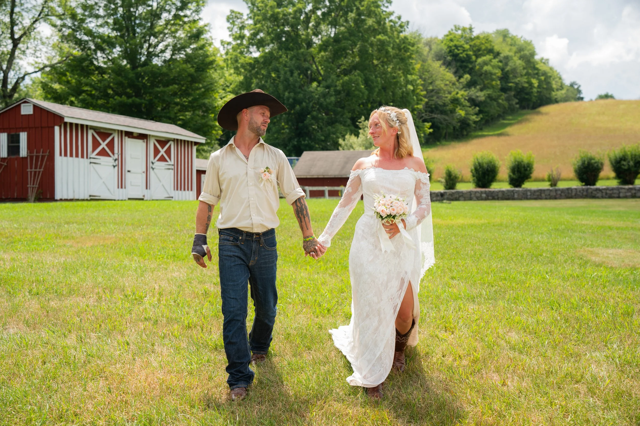 Bride in white lace dress and cowboy boots holds bouquet, groom in cowboy hat and jeans, walking hand in hand on a grassy field with rustic barn background.