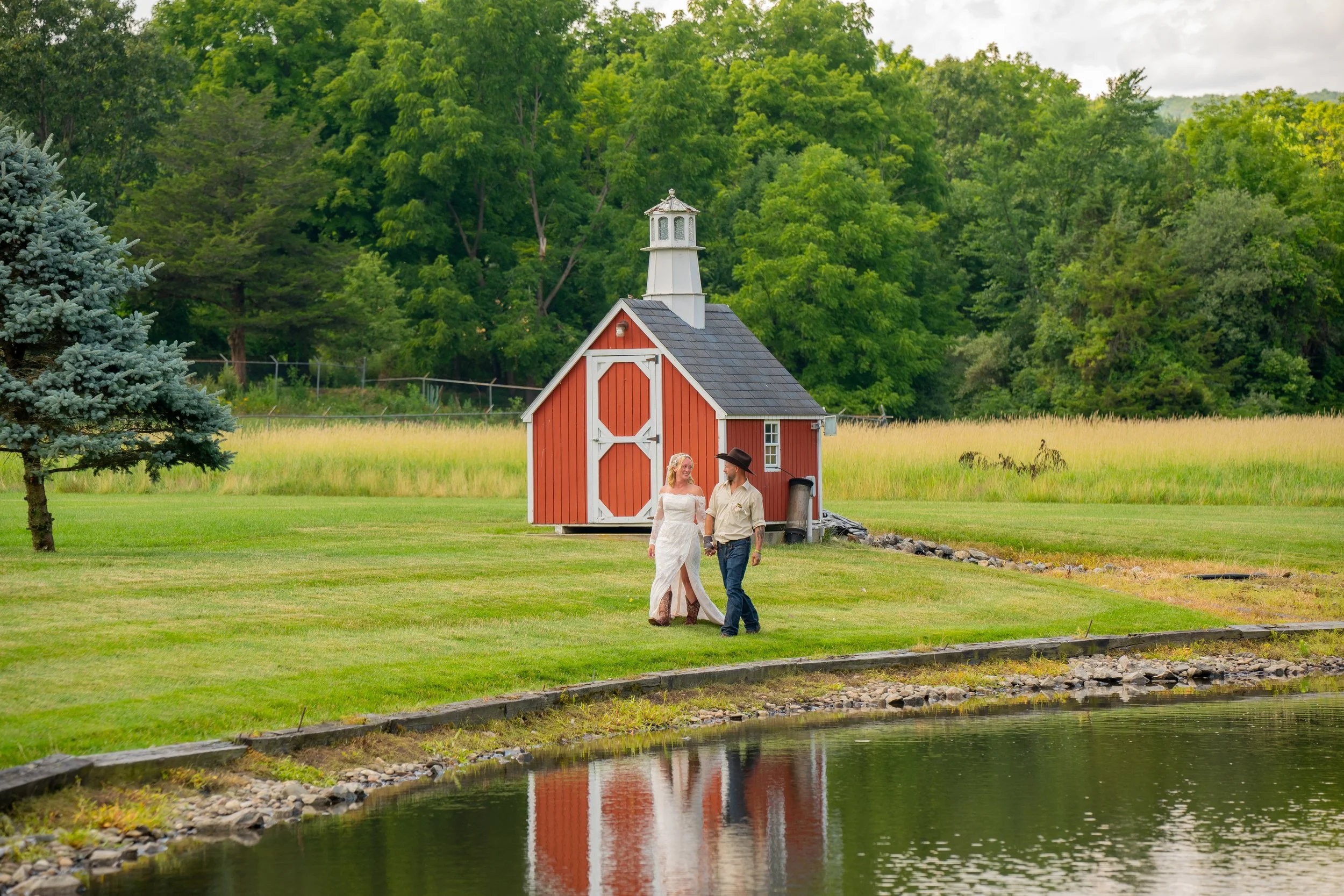 Couple walking near a barn by a pond in a rural landscape with trees and grass.
