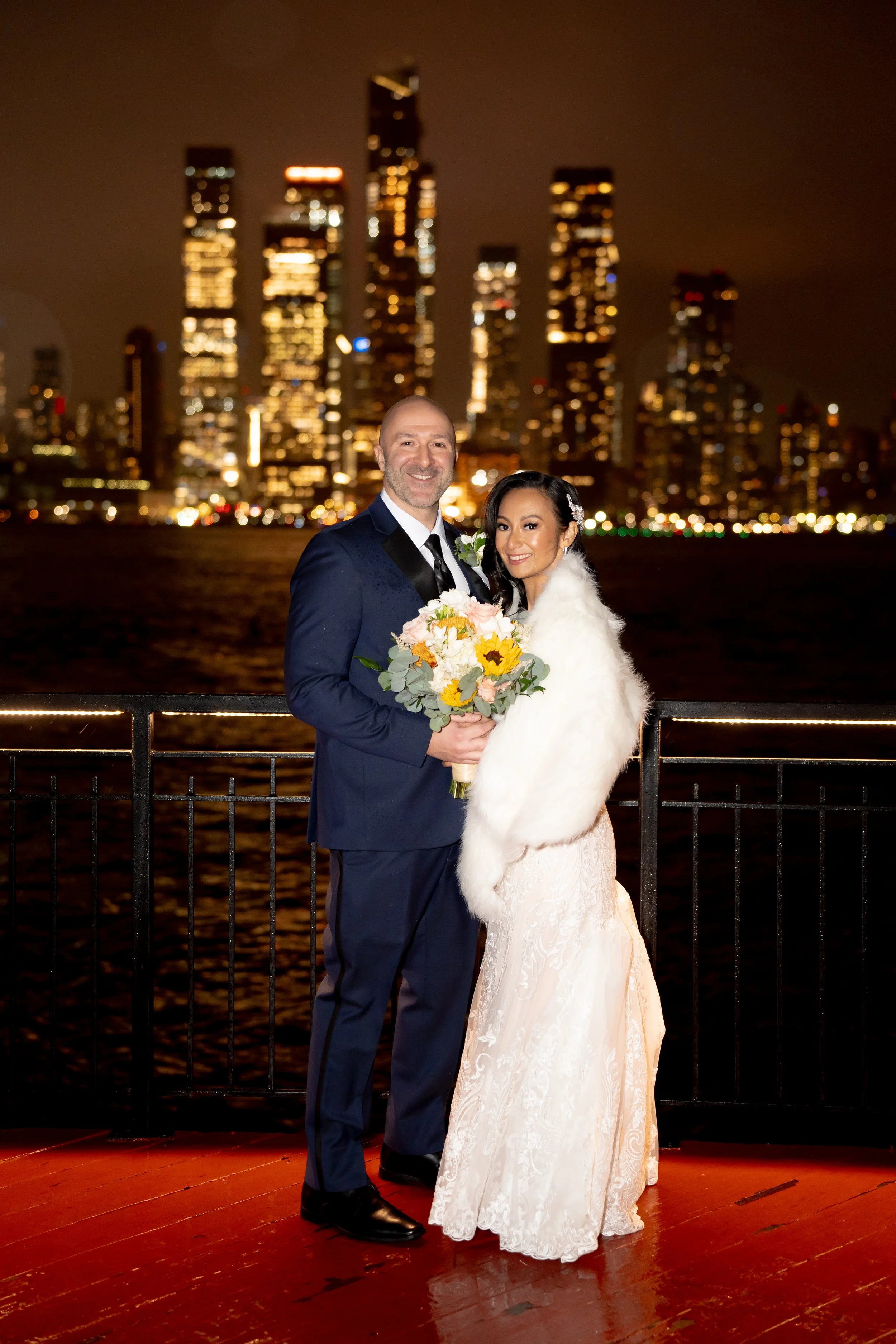 Couple in wedding attire posing at night with lit city skyline in the background.