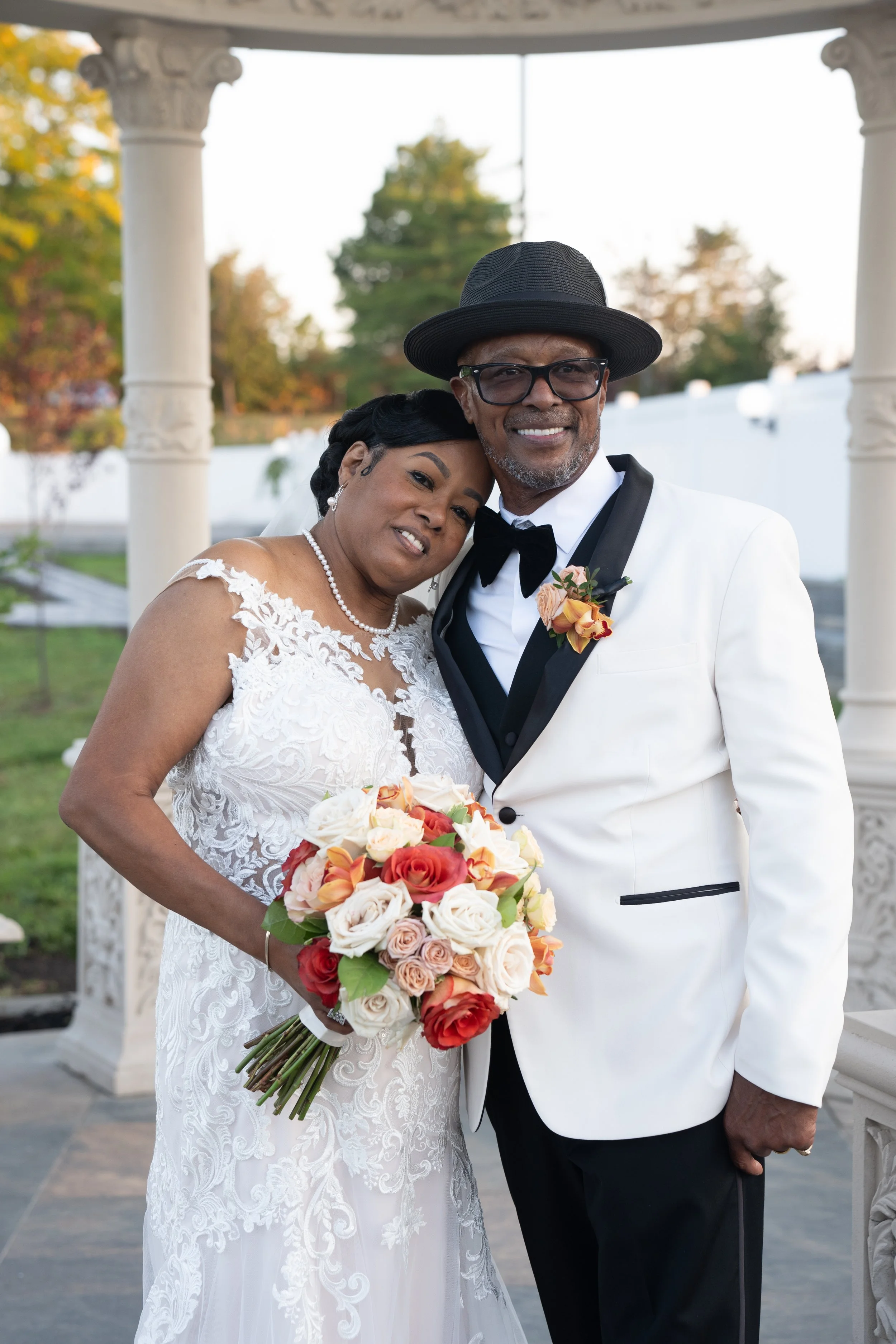 Bride and groom smiling, bride holding colorful bouquet, groom in white suit, outdoor setting.