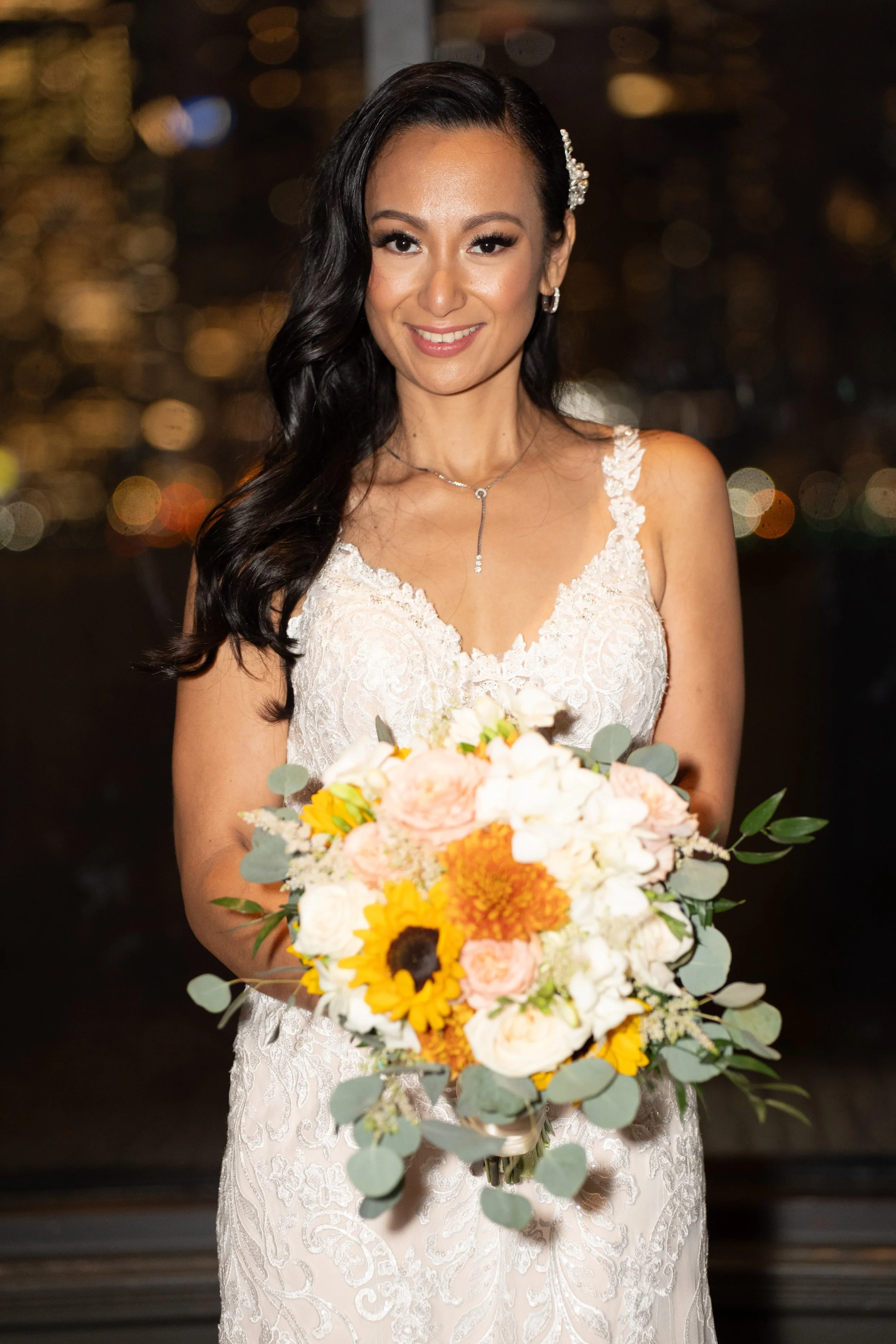 Bride in lace wedding dress holding a bouquet with sunflowers, roses, and greenery.