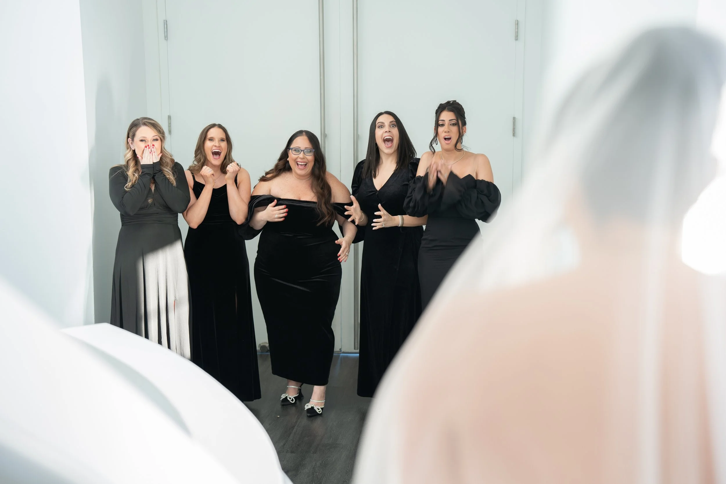 Five women in black dresses reacting to seeing a bride in a wedding dress with a veil.