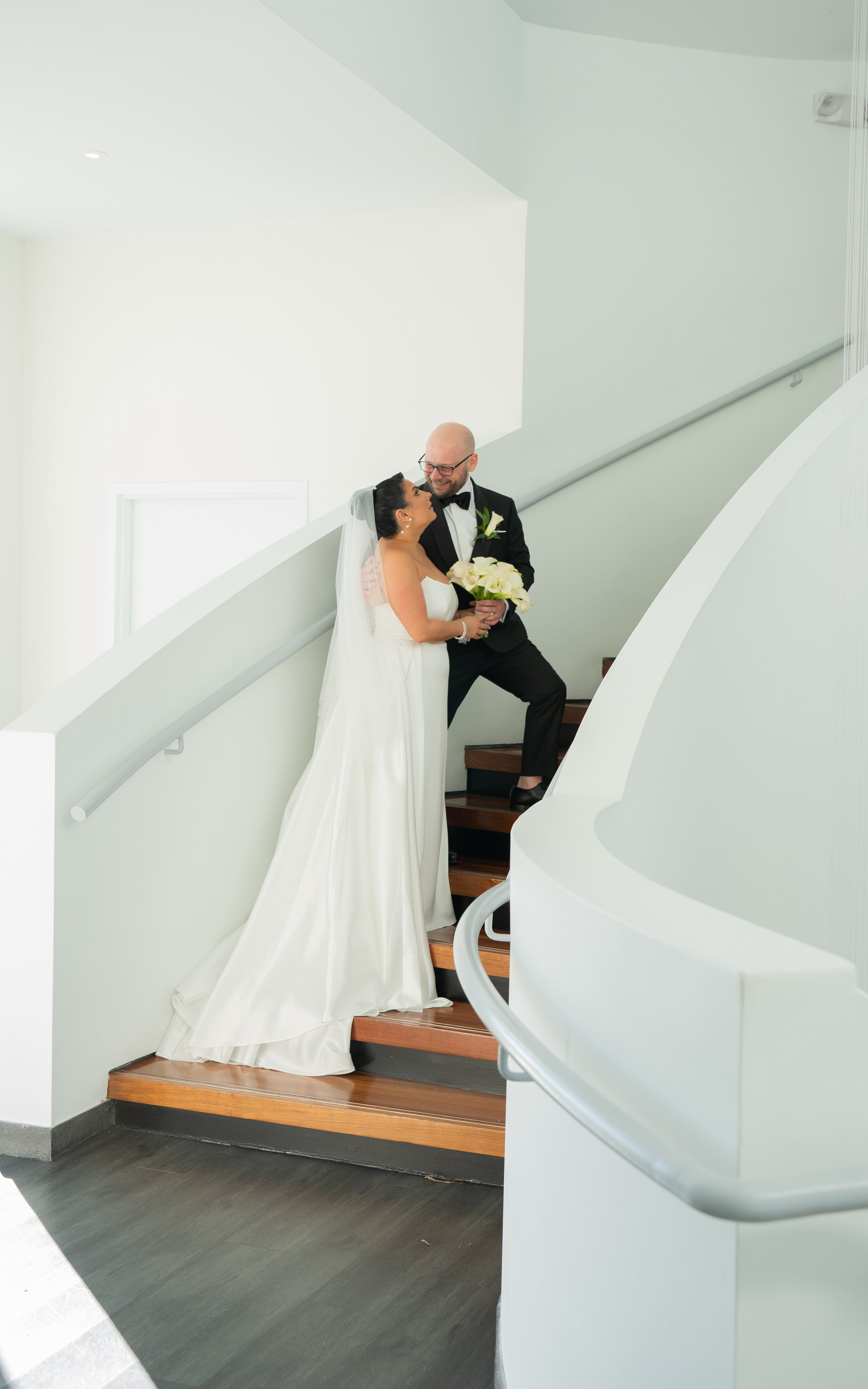 Bride and groom embracing on staircase, bride in white gown holding bouquet, elegant wedding scene.