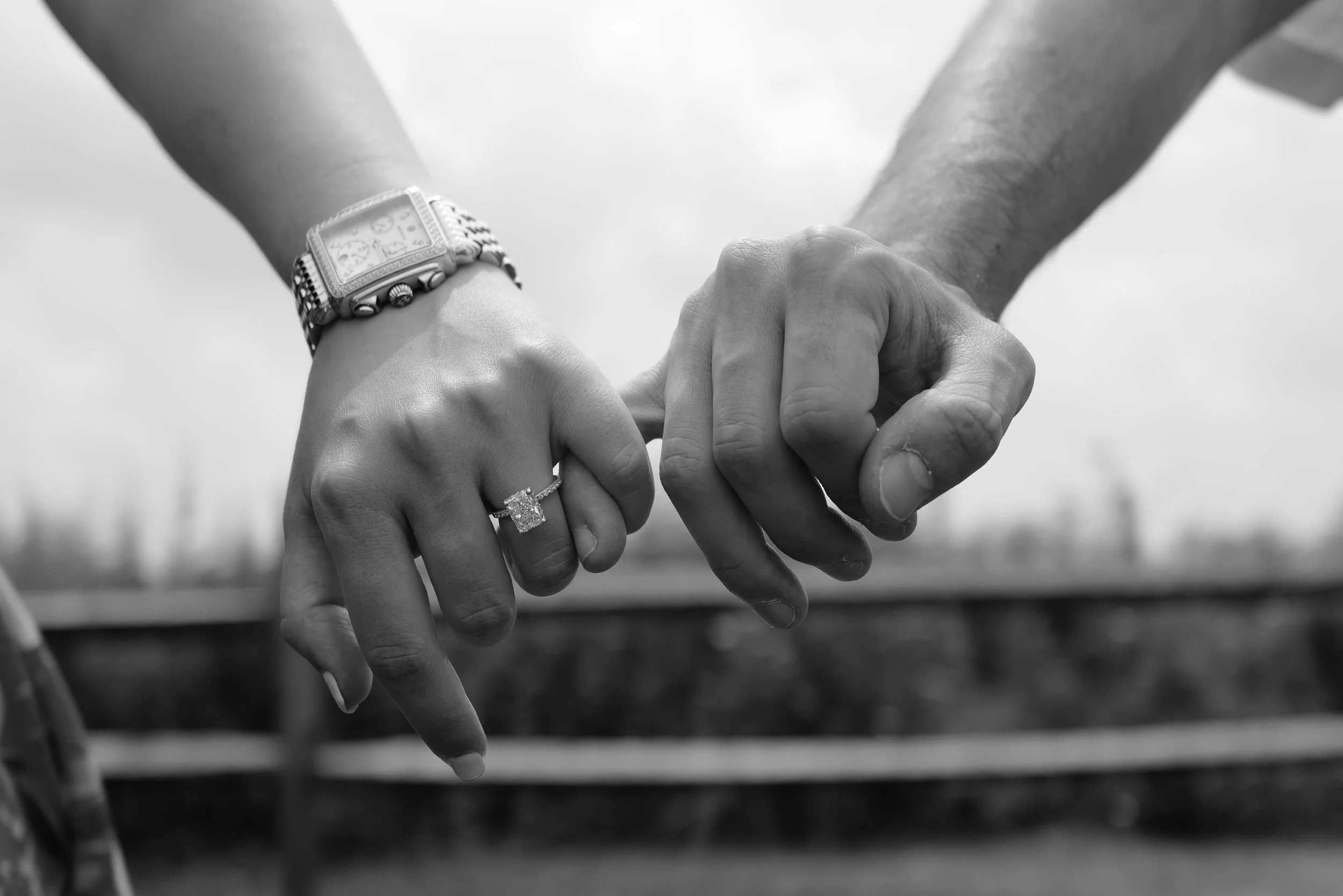 Black and white image of two people holding hands with focus on a woman's hand wearing a watch and an engagement ring.