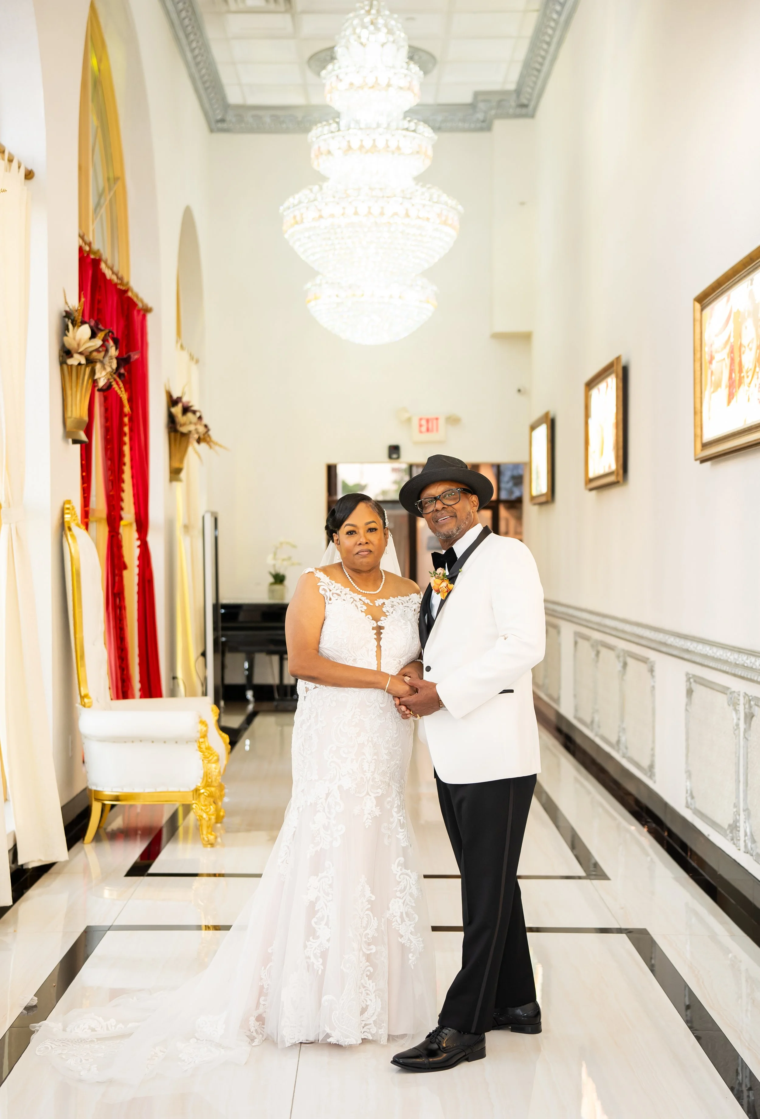 Elegant couple posed in a luxurious hallway with chandeliers, wearing formal wedding attire; the woman in a white bridal gown and the man in a white jacket and black hat.