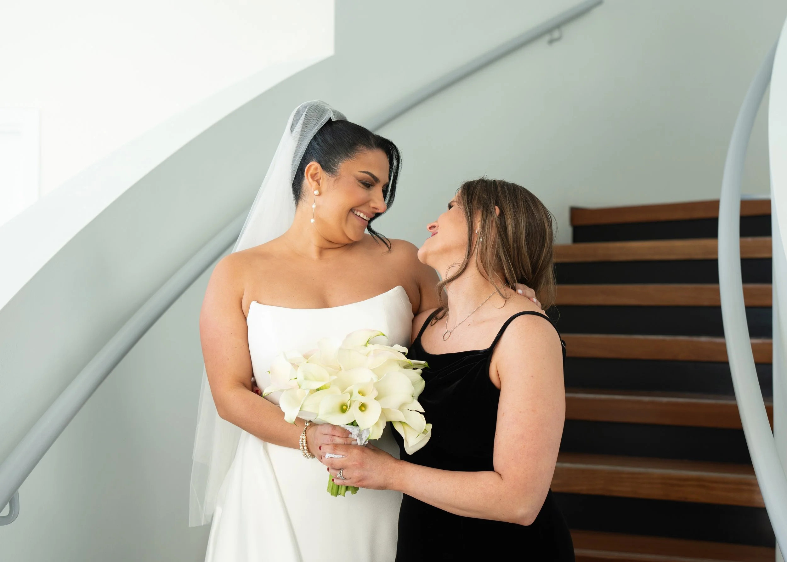 Bride in white dress holding flowers, smiling with another woman in a black dress, standing by a staircase.
