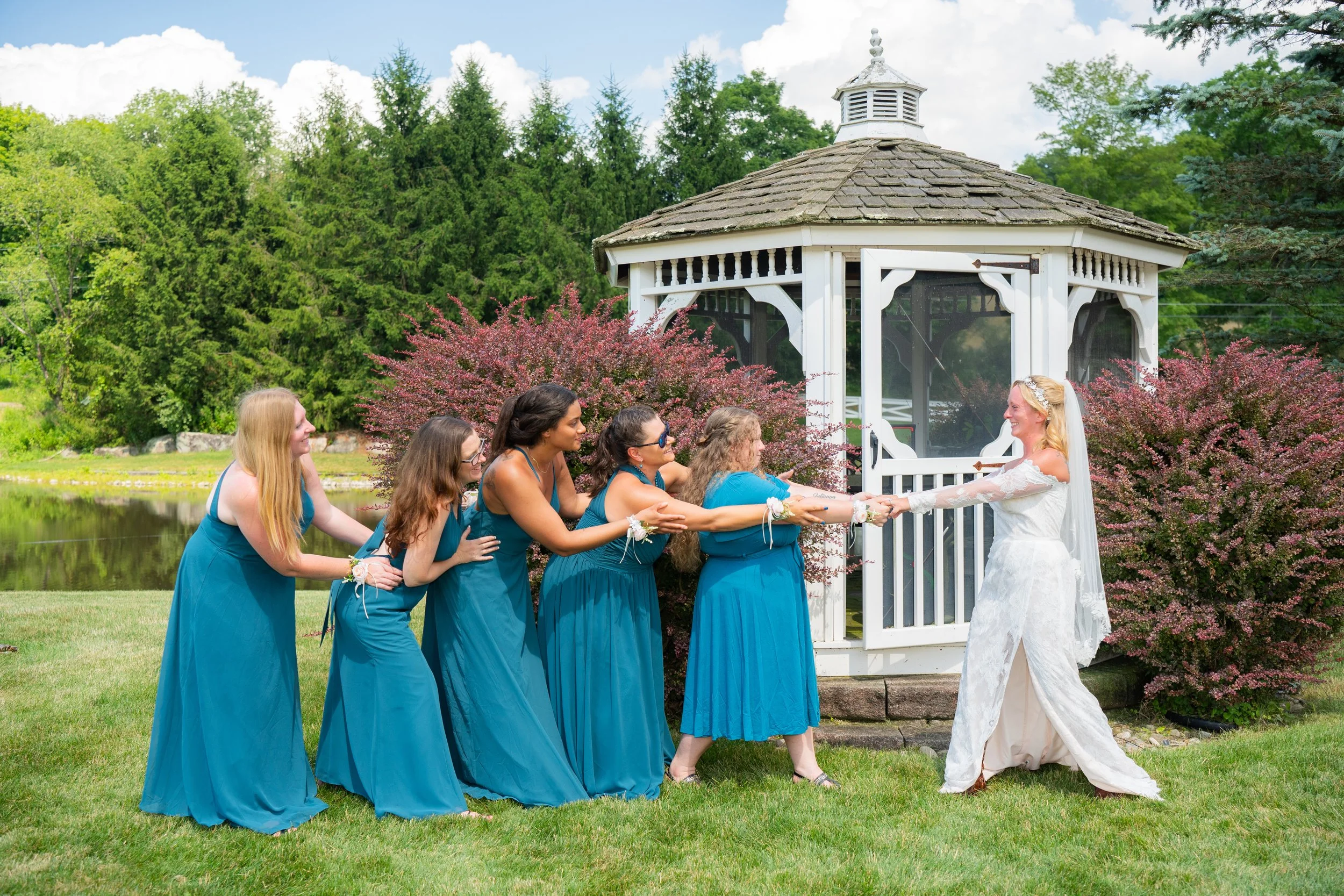 Bridesmaids in blue dresses playing tug-of-war with the bride in front of a gazebo, outdoor wedding setting.