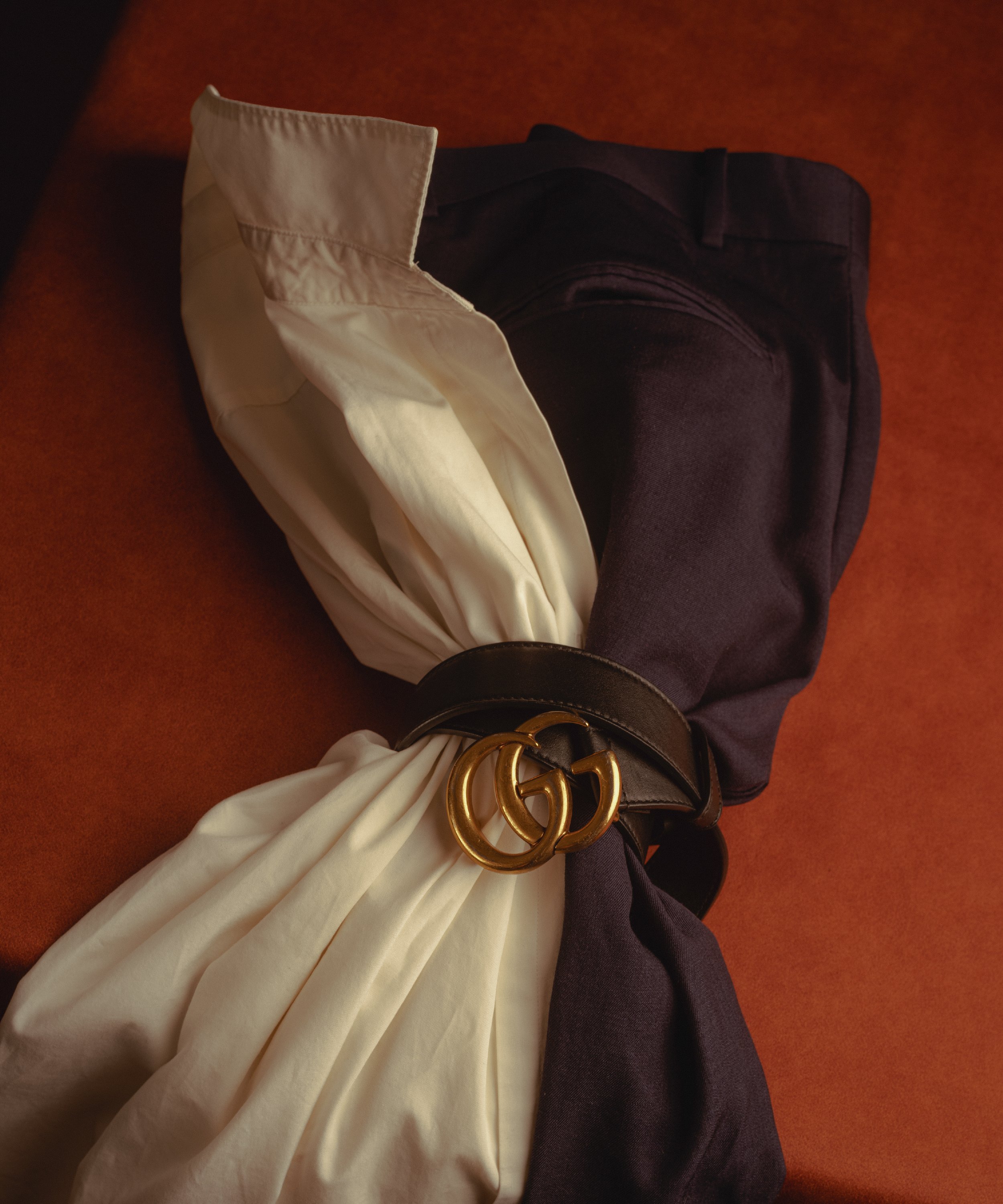Folded pants and shirt with a black belt and gold buckle on a brown surface.