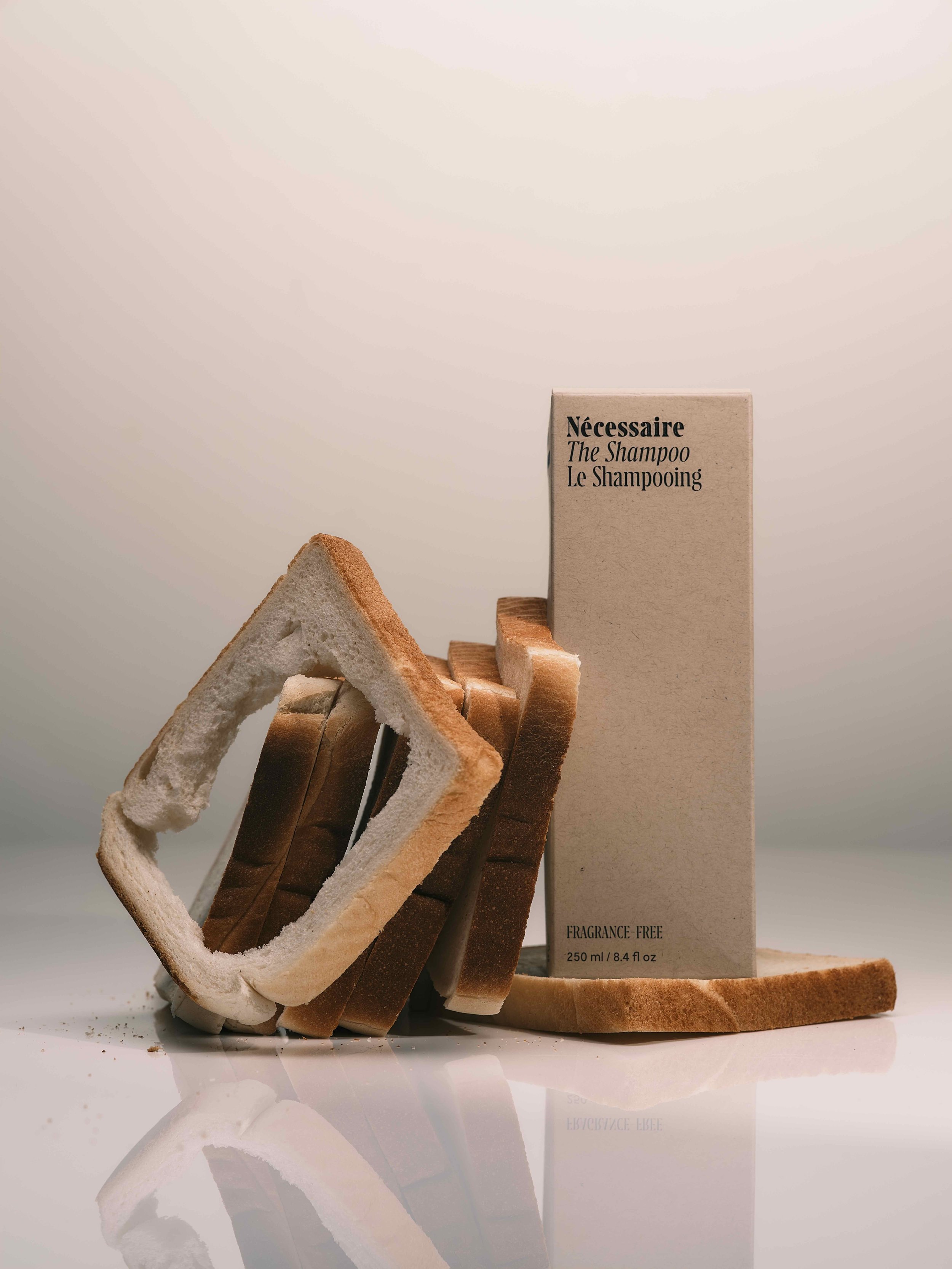 A bottle of Nécessaire The Shampoo, labeled as fragrance-free, surrounded by slices of bread, including one with the crust removed and hollowed out. The setting is a neutral background.