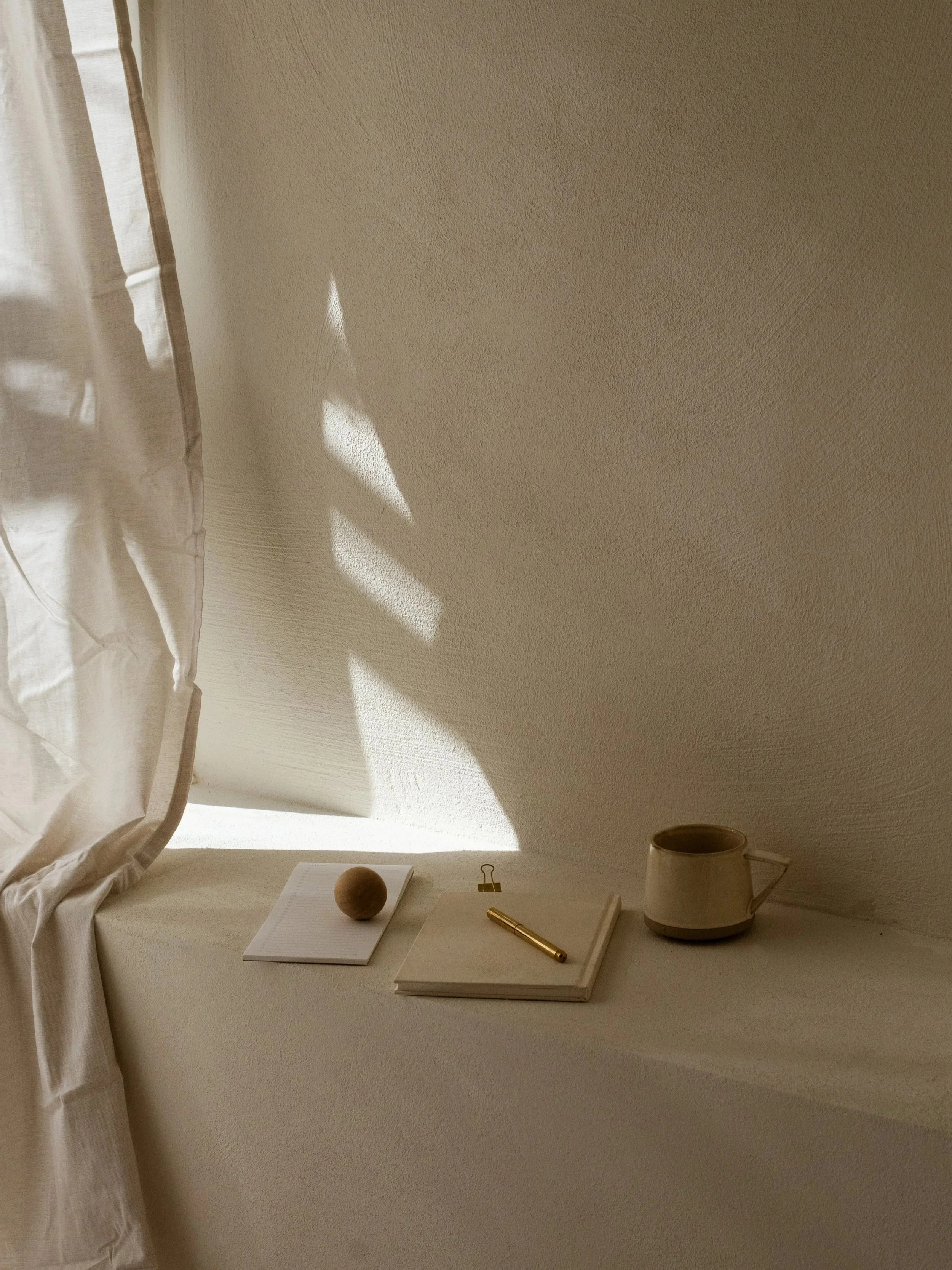 A beige shelf with a white notebook, a small notebook with a gold pen, a clip, a mug, and a round wooden object, with sunlight creating shadow patterns on the wall.