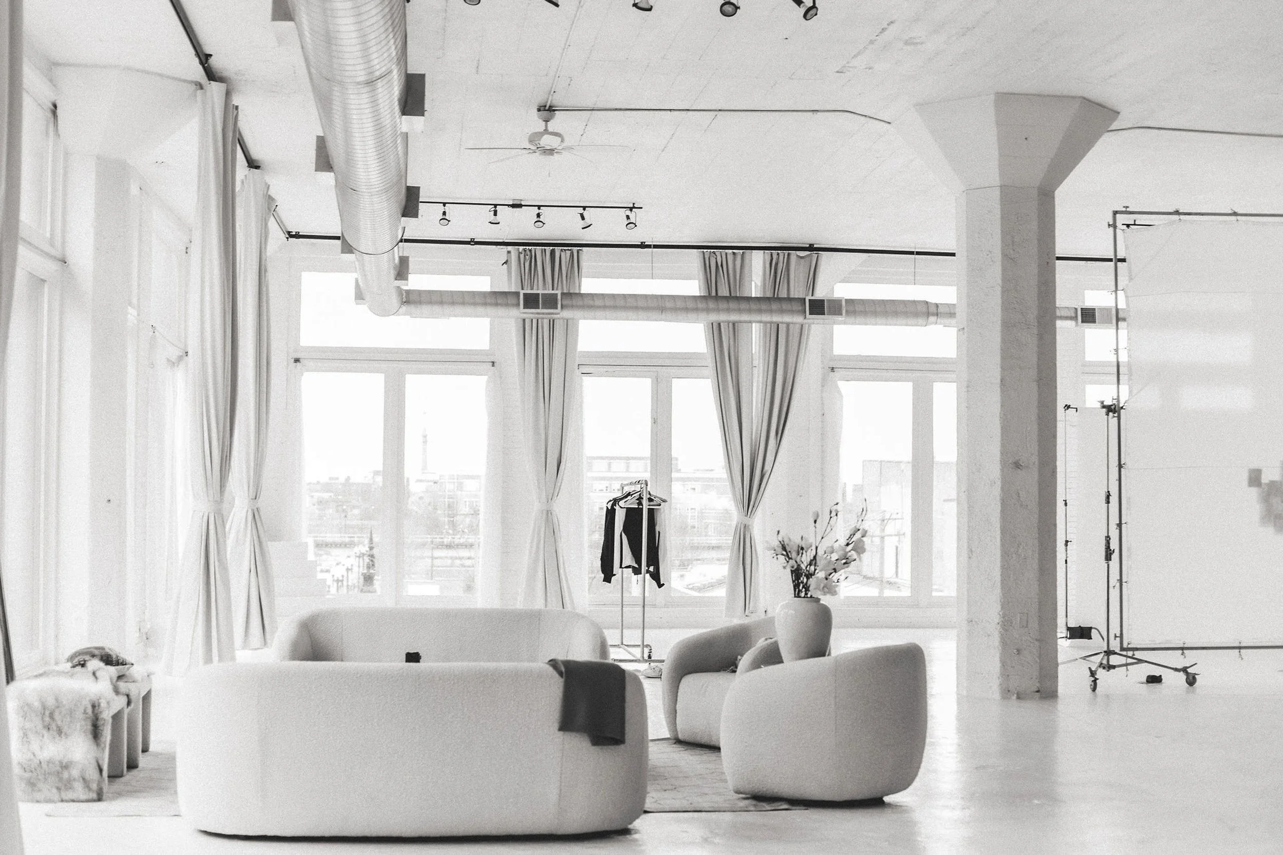 A spacious, sunlit room with large windows, white curtains, and modern furniture including sofas, a vase with flowers, a clothing rack with garments, and studio lighting equipment.