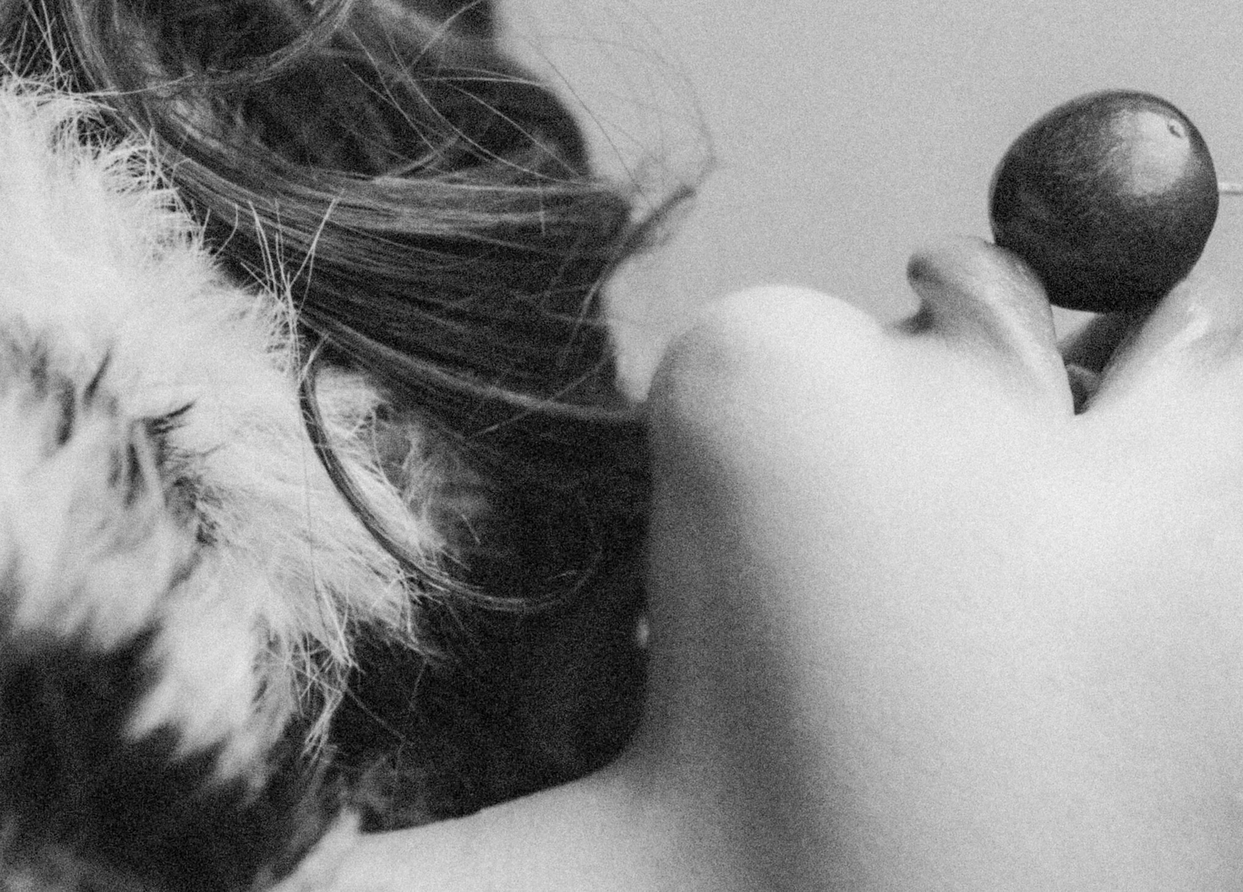 A person lying down and holding a round object, possibly a ball or a fruit, with their hand. The person's face is partially visible, and they have long hair. The image is in black and white.