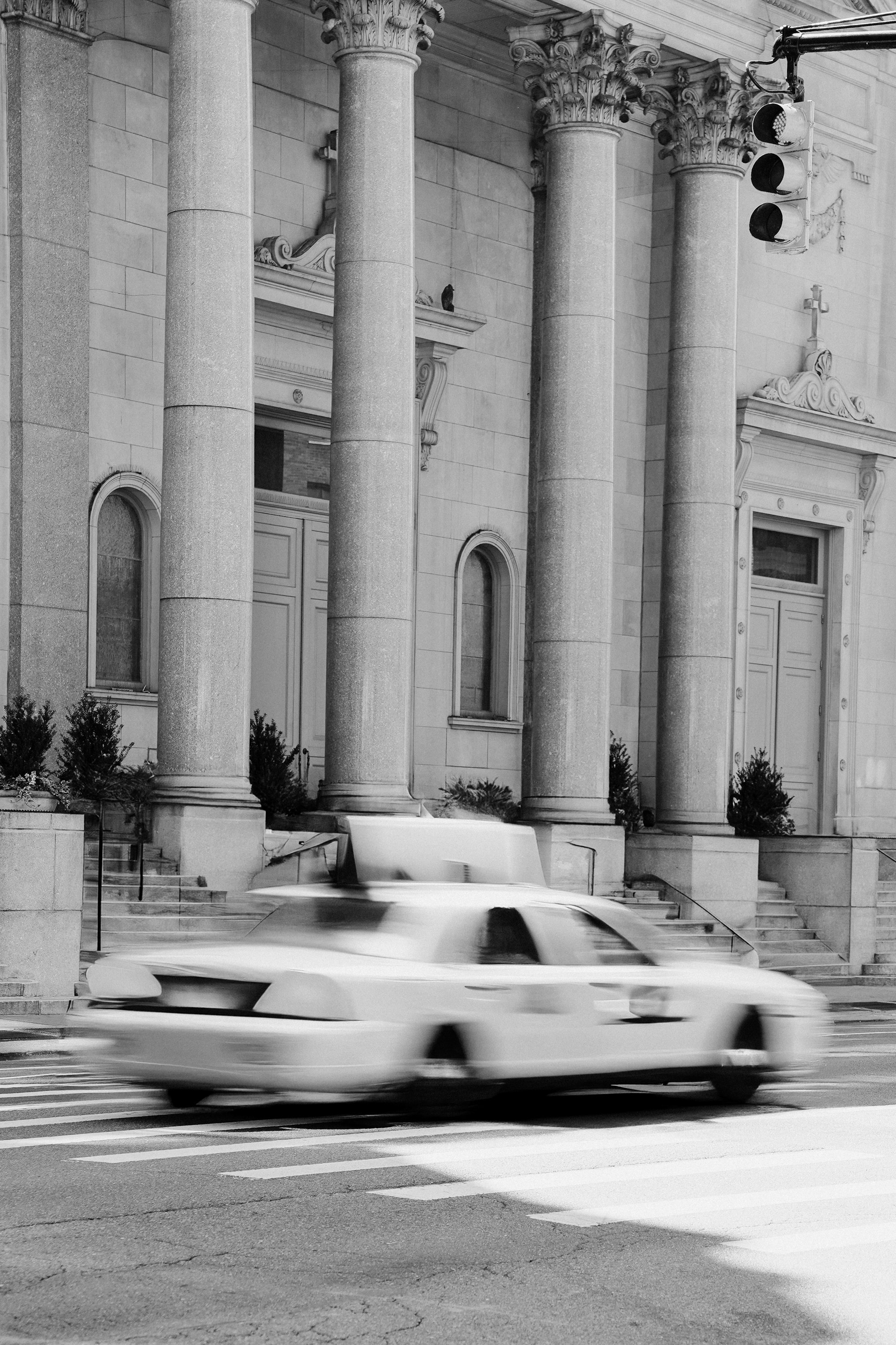 A black and white photo of a city street with a blurred white police car driving past a building with large columns, stairs, and small bushes along the sidewalk. A traffic light is visible in the upper right corner.