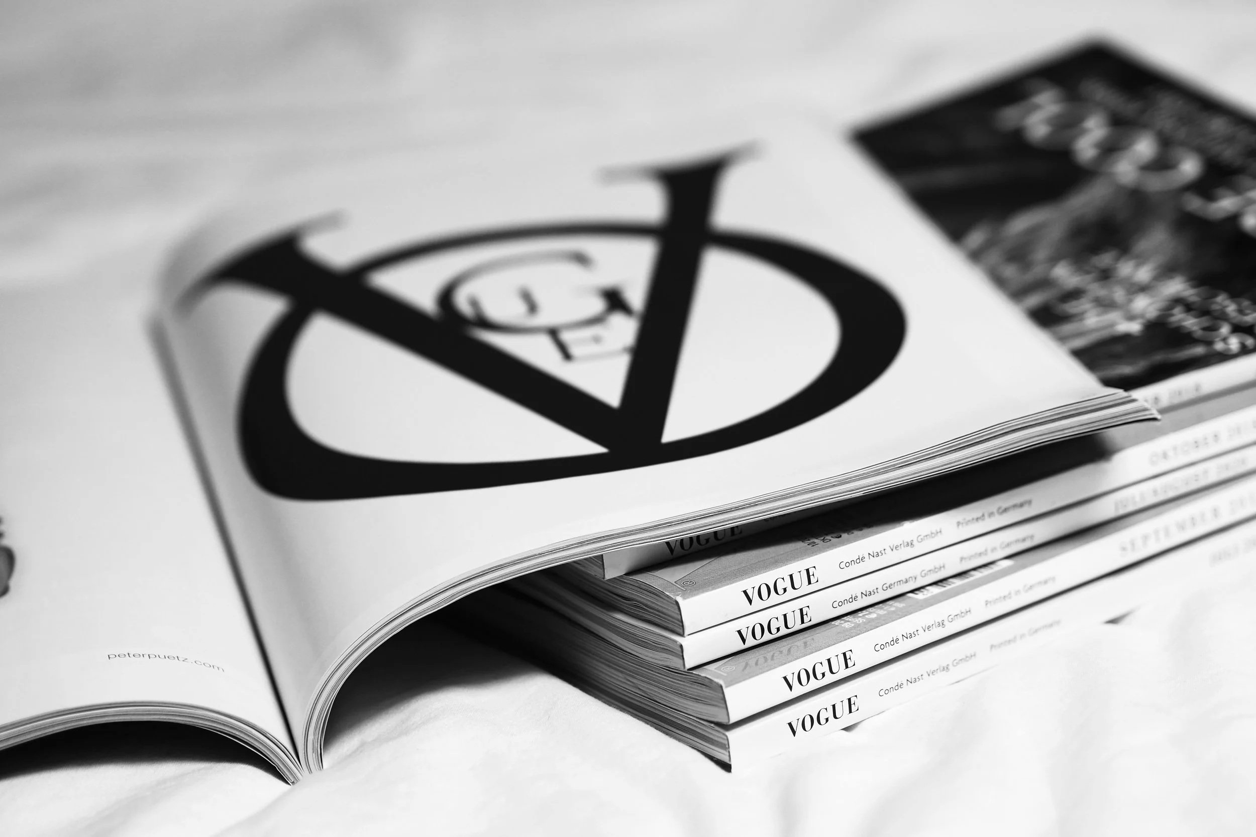 Black and white magazine pages, with the VOGUE logo on the spine of three stacked magazines and an open magazine showing a large grayscale logo on the page.