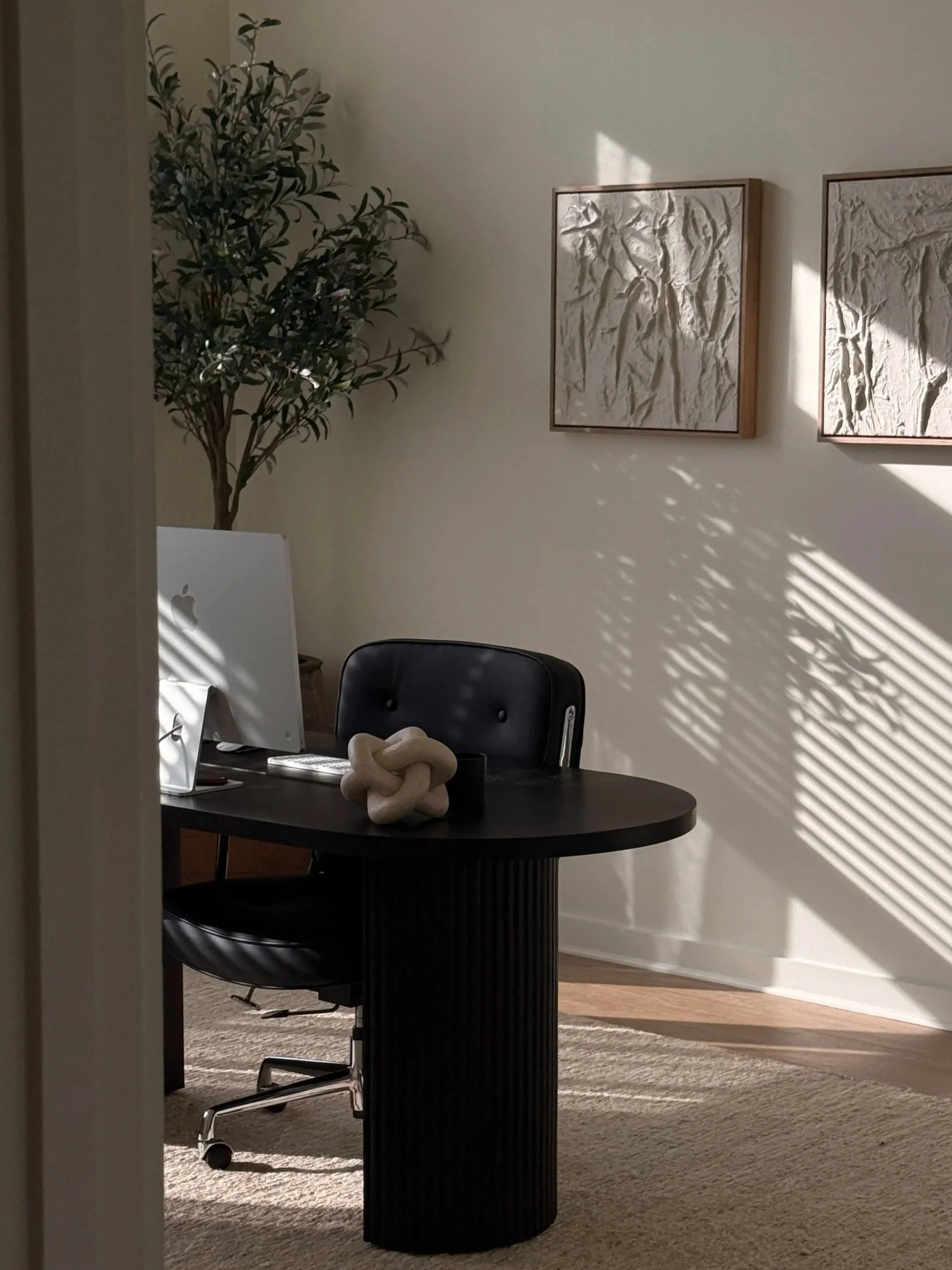 An office desk with a computer and decorative object, in a room with artwork on the wall and sunlight casting shadows.