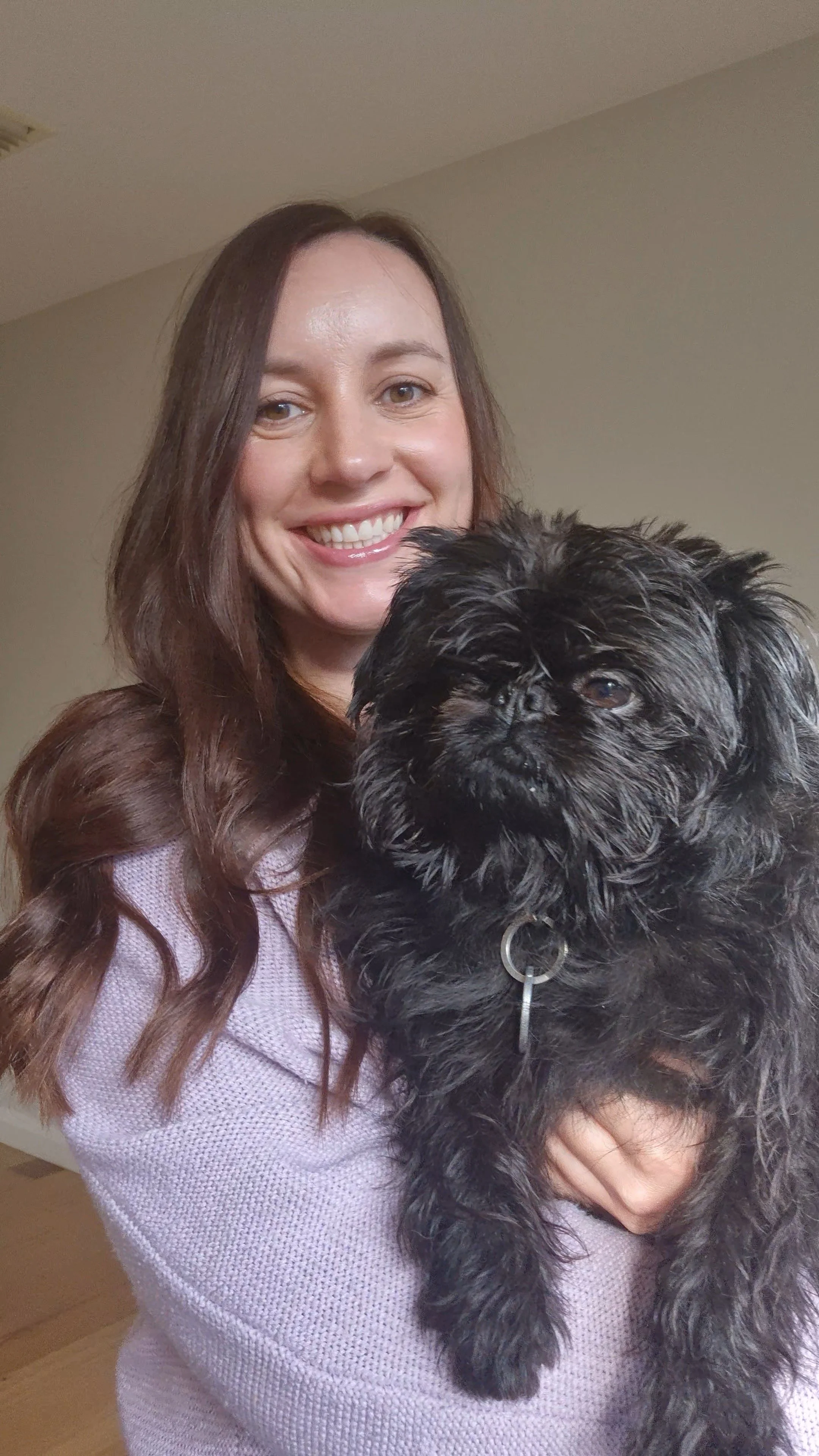 A smiling woman with long brown hair holding a small black dog with curly fur in front of a beige wall.