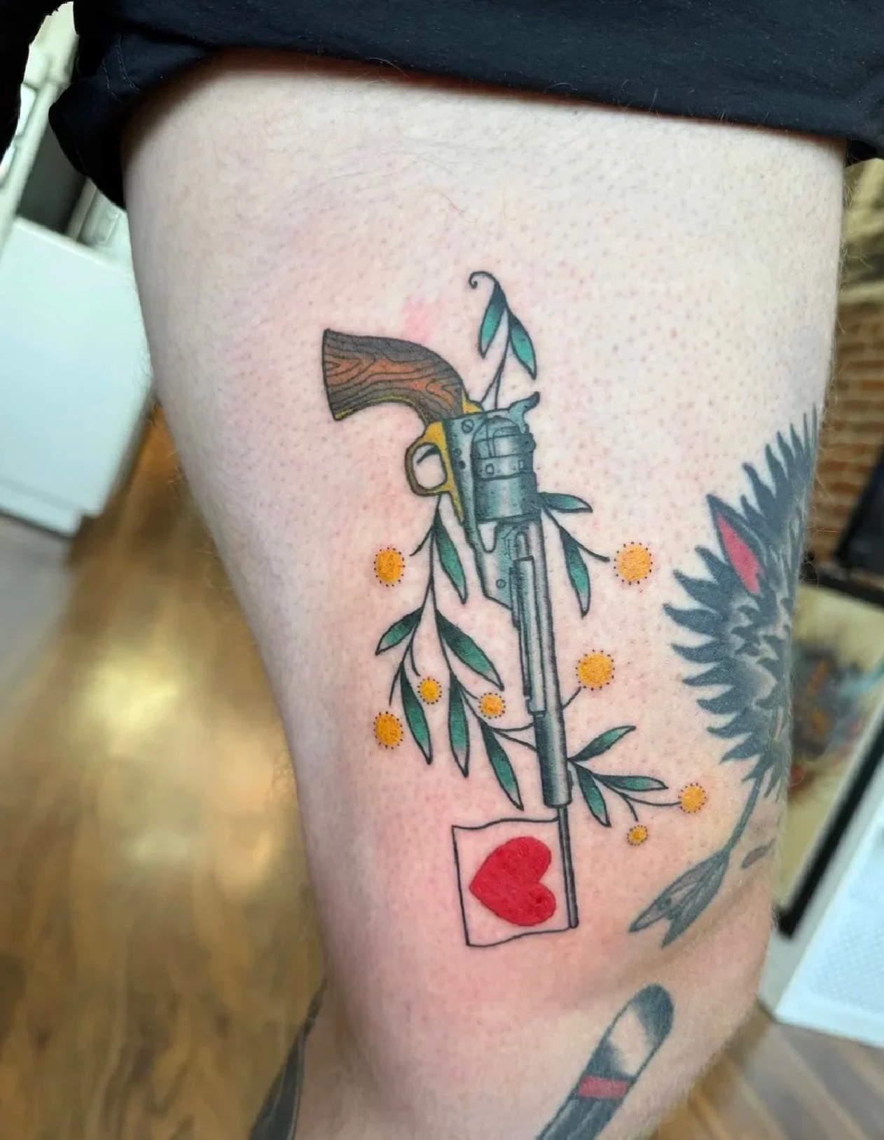A classic revolver softened with florals and topped off with an adorable heart flag tattooed by @charliedarling_tattoo ✨ We LOVED the balance of bold lines with those tiny pops of color! 🩷 #revolver #cute #tattoo #artist #junipertattoo