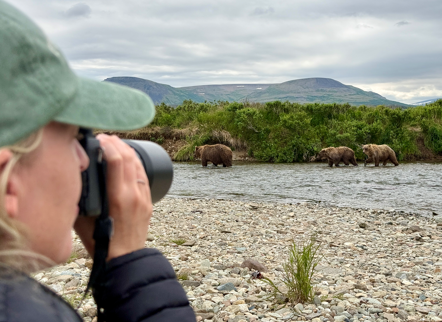 A blonde woman in a green hat and black jacket holding a camera taking a photo of bears in a river with a mountain and cloudy sky in the background.