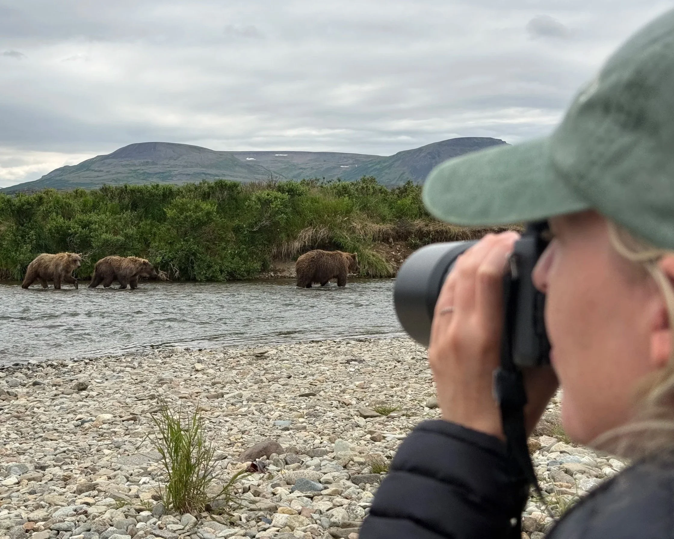 A blonde woman in a green hat and black jacket holding a camera taking a photo of bears in a river with a mountain and cloudy sky in the background.