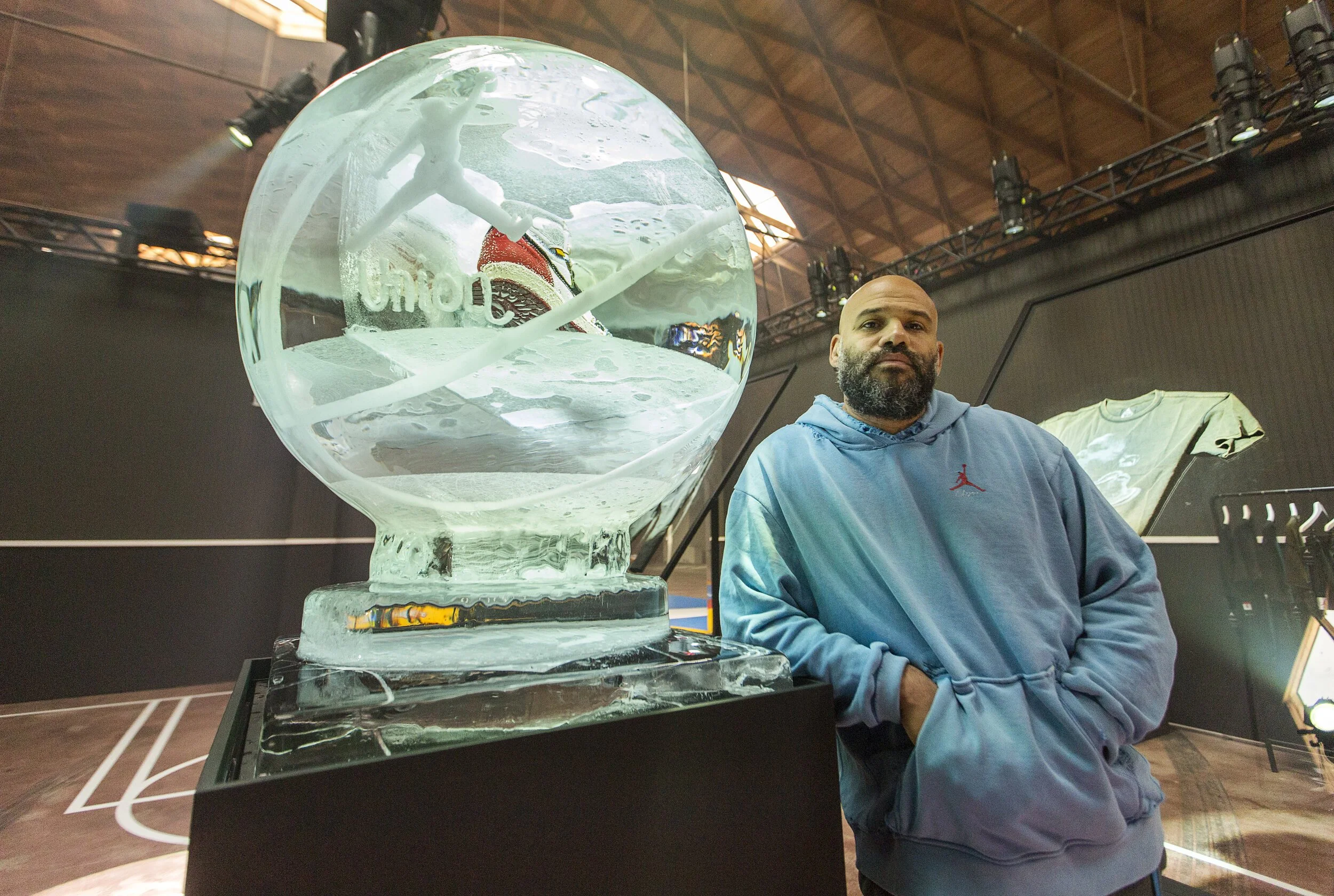 A man with a beard in a blue hoodie stands next to an ice sculpture of a basketball with a glass ball design, inside a basketball court with display screens and clothing in the background.