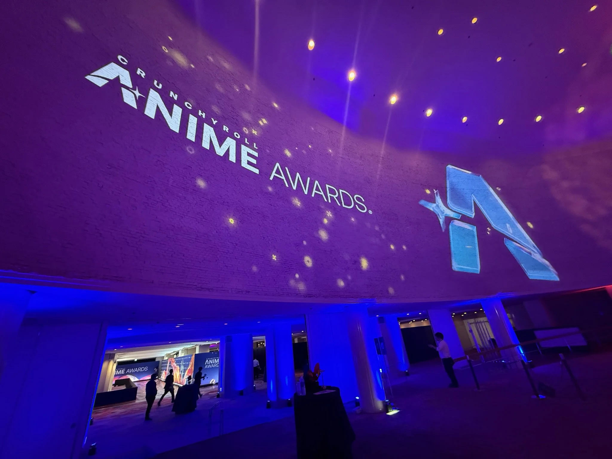 Large screen displays 'Crunchyroll Animae Awards' on a purple background with glowing lights, amid a dimly lit event space with a few people walking around.