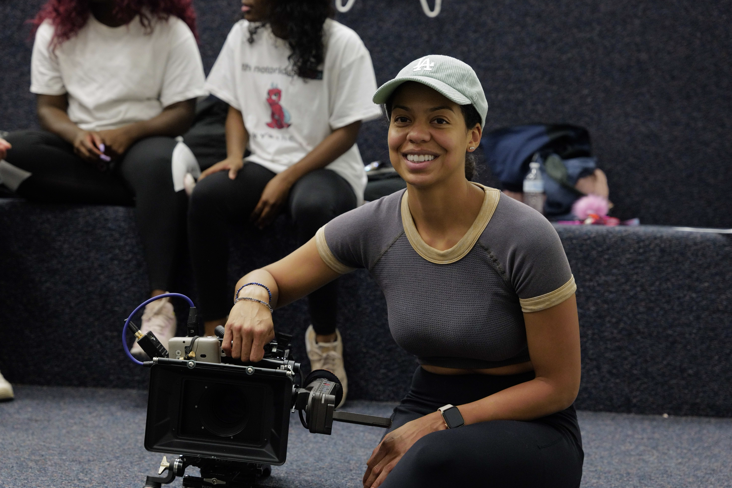 Woman smiling while operating a professional video camera, seated on carpeted floor in a room with dark textured walls, with two women sitting on steps behind her.