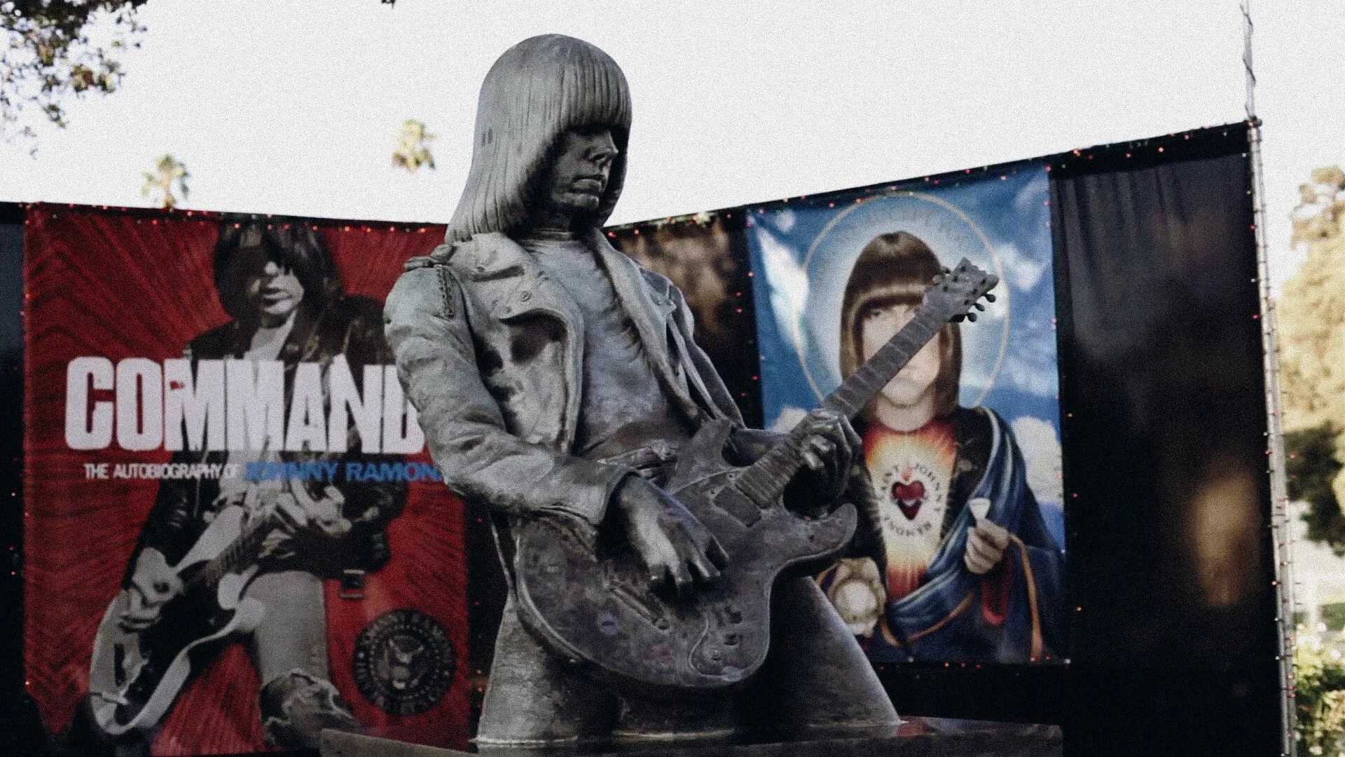 Statue of a young person with long hair playing an electric guitar in front of a promotional poster for the TV series 'The Commandos' with the character 'Andy Ramsey' on it.