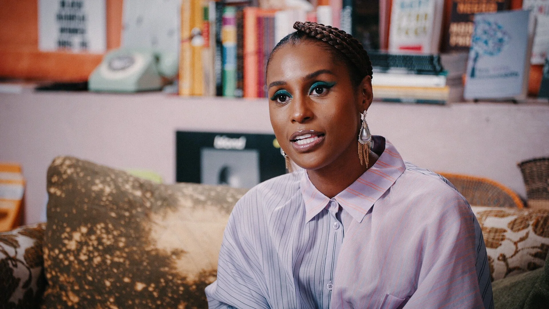 A woman with braided hair wearing a striped button-up shirt, colorful makeup, and long earrings, sitting on a sofa in a room with shelves of books and a vintage rotary phone in the background.
