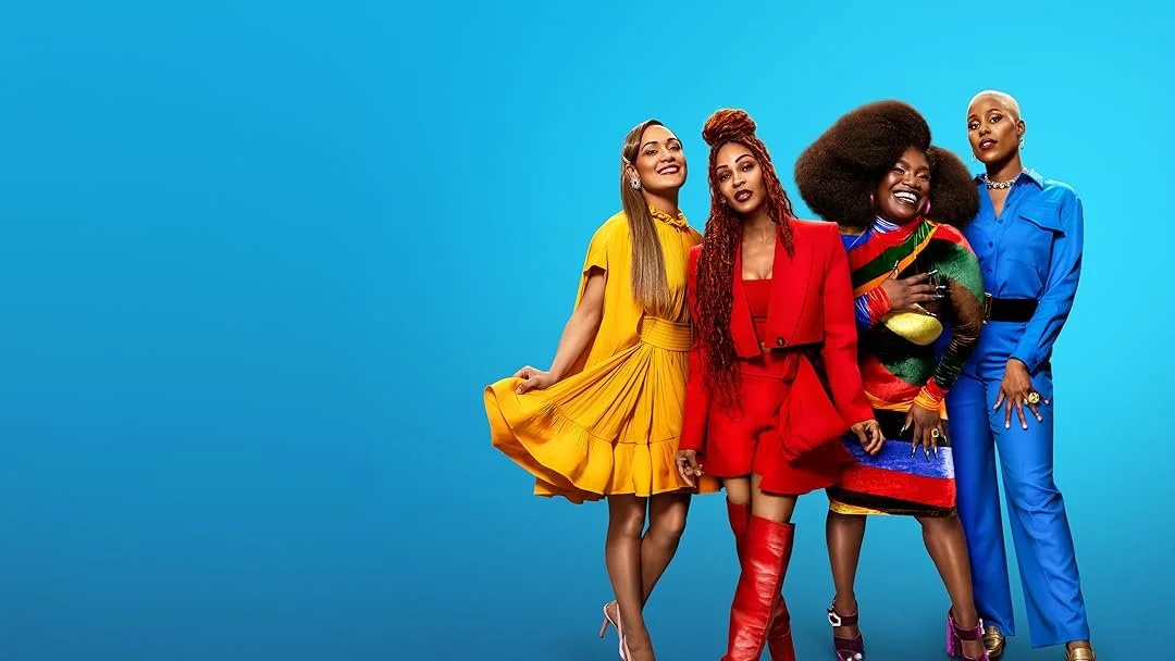 Five women standing together against a light blue background, dressed in colorful fashionable outfits, smiling and posing for the camera.