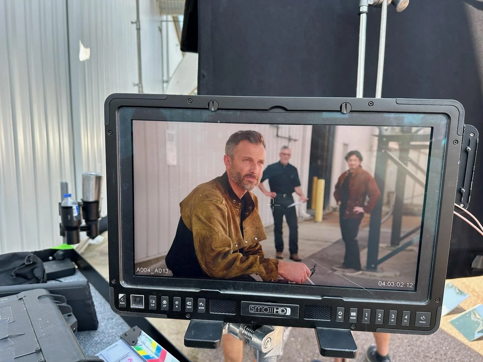 A video camera monitor shows three men in a warehouse setting during a film shoot; the owner of the company is sitting close to the camera, with two others standing in the background.