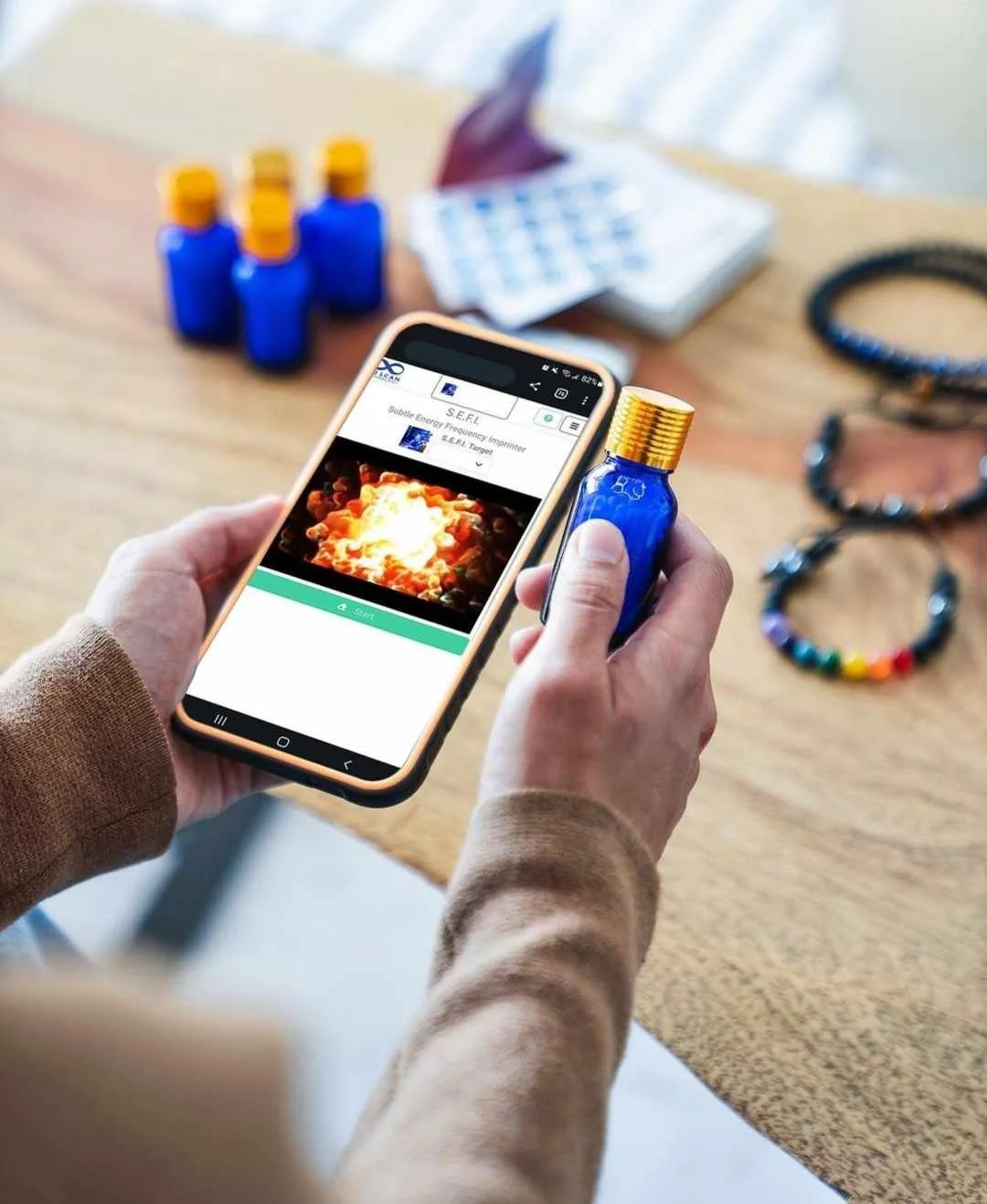 Person holding a smartphone with an image of an explosion on the screen. The table in the background has several small blue bottles with yellow caps, a few jewelry pieces, and a blurred blister pack. AO SEFI technology