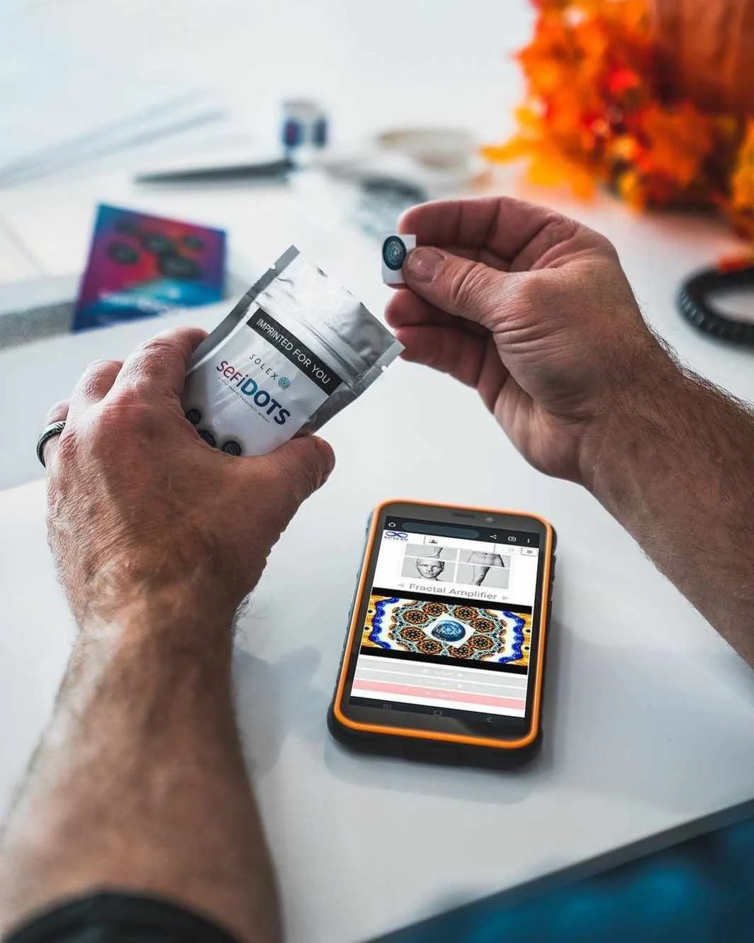 A person holding a package of Dot stickers and peeling one off, with a smartphone displaying an image of a colorful pattern and text about fractal amplifiers SEFI on a white desk.