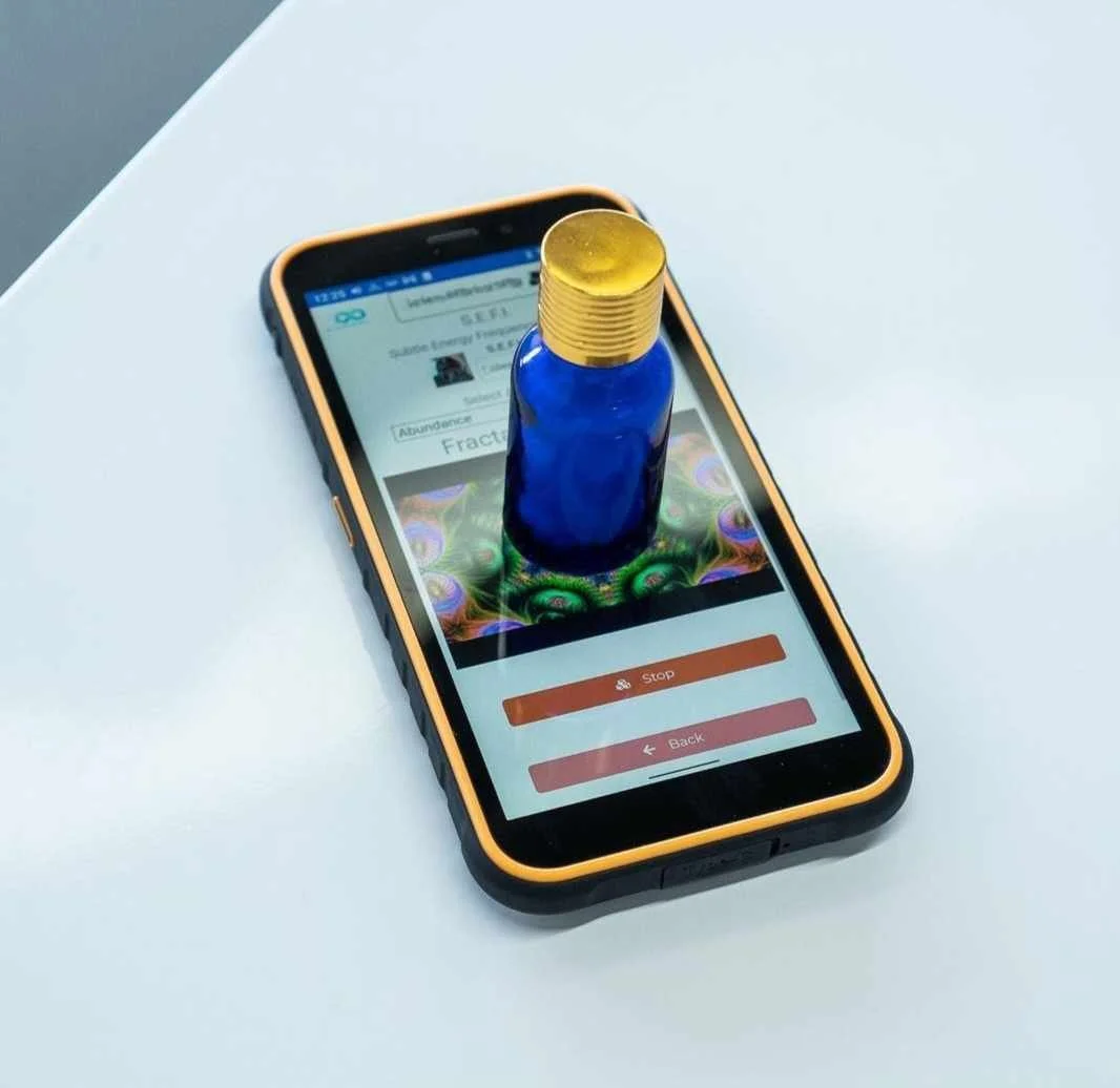 A smartphone on a white surface displays an app with a blue bottle on top of it. The app shows a colorful, frequency generator from AO scan