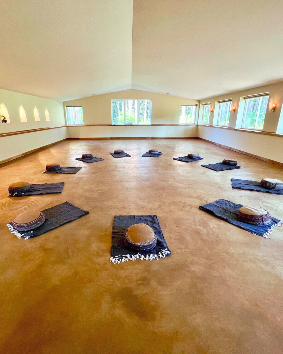 An empty meditation or yoga room with 12 black mats and brown round cushions placed on top, arranged in a circle. The room has large windows on two sides, beige walls, and a polished wooden floor.