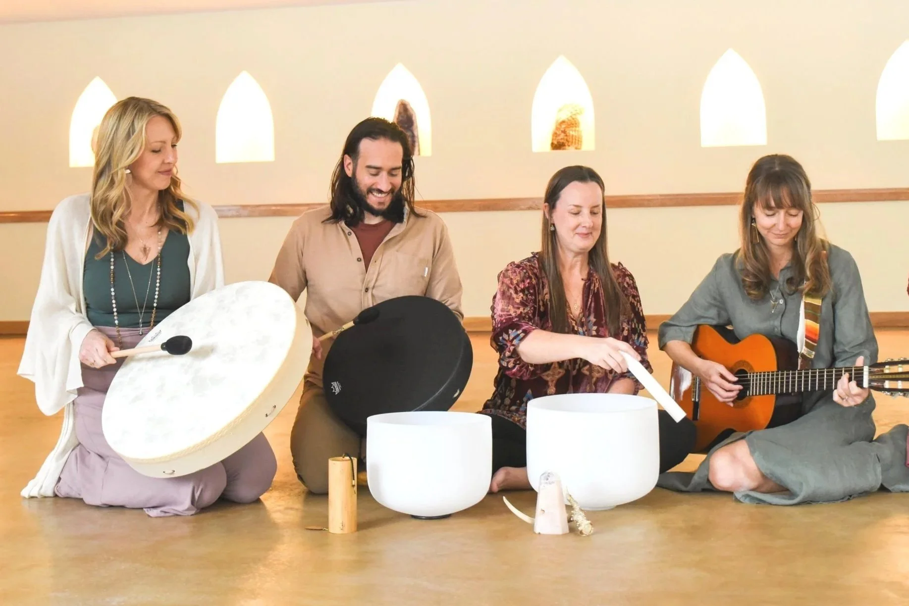 Group of six people sitting on the floor in a semi-circle, playing musical instruments in a room with arched windows and wooden accents.