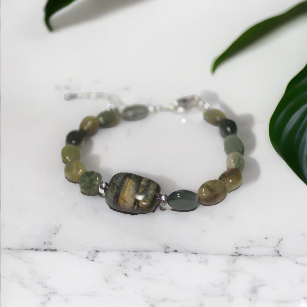 Beaded bracelet featuring a large central stone, surrounded by smaller green, black, and brown beads, placed on a white marble surface with green leaves nearby.
