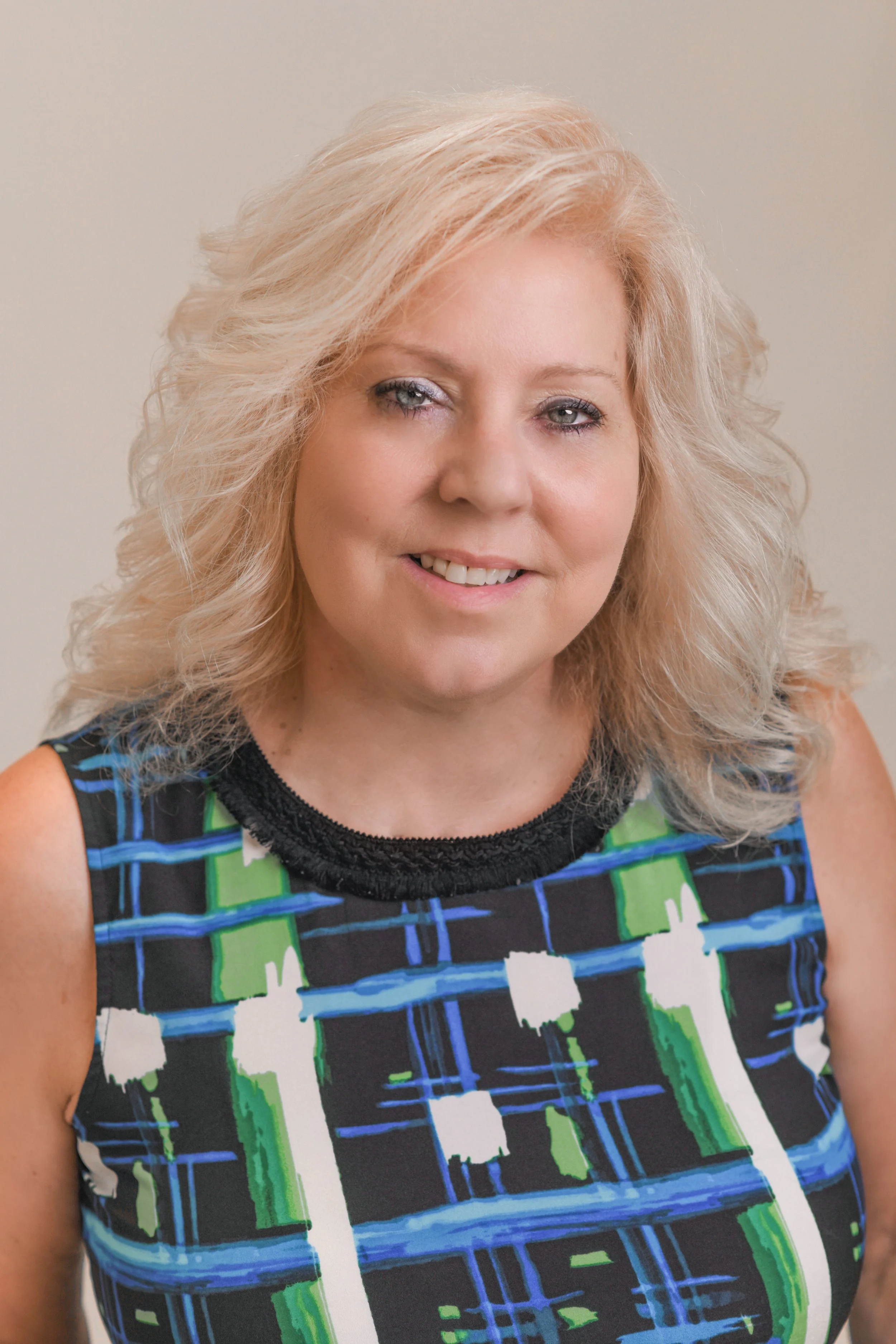 A woman with shoulder-length blonde hair wearing a sleeveless dress with a colorful abstract pattern in green, blue, black, and white.